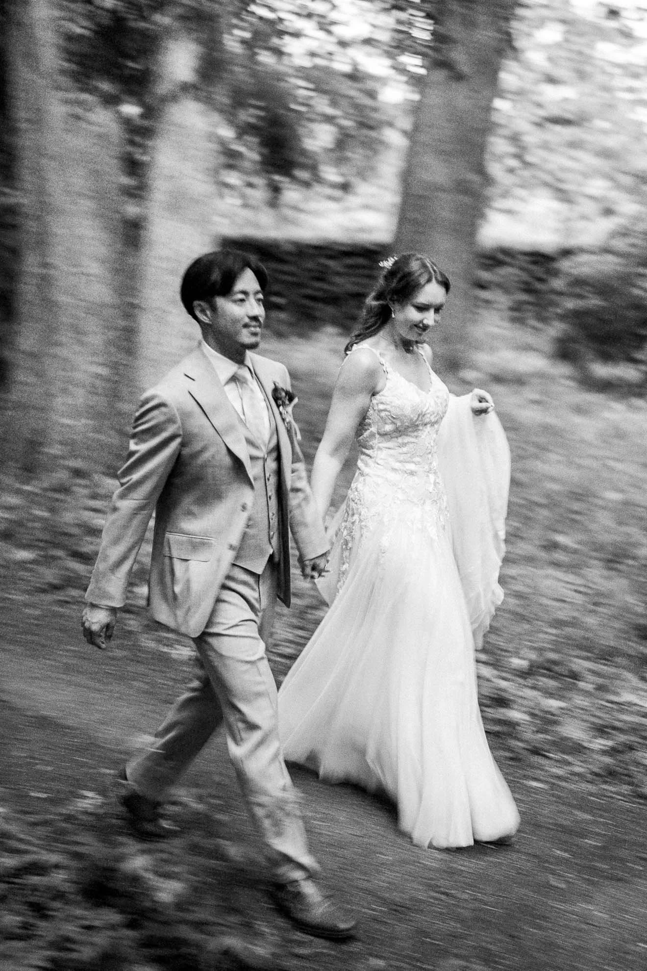 A black-and-white photo of a bride and groom walking hand in hand outdoors, with trees in the background.