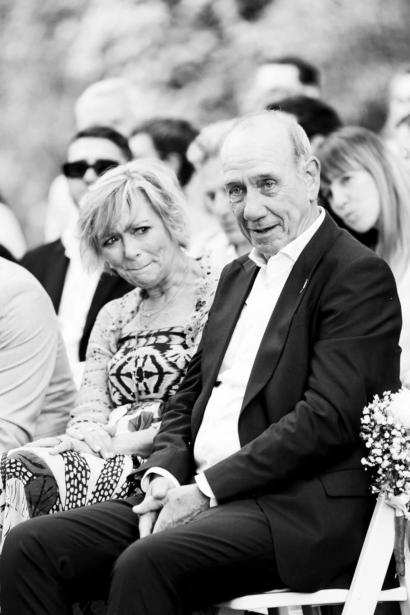 A group of people sitting in a row at an outdoor event. The focus is on a man in a suit and a woman sitting next to him. The woman has short, light-colored hair and appears to be upset or displeased. The man has a serious expression. Other individual