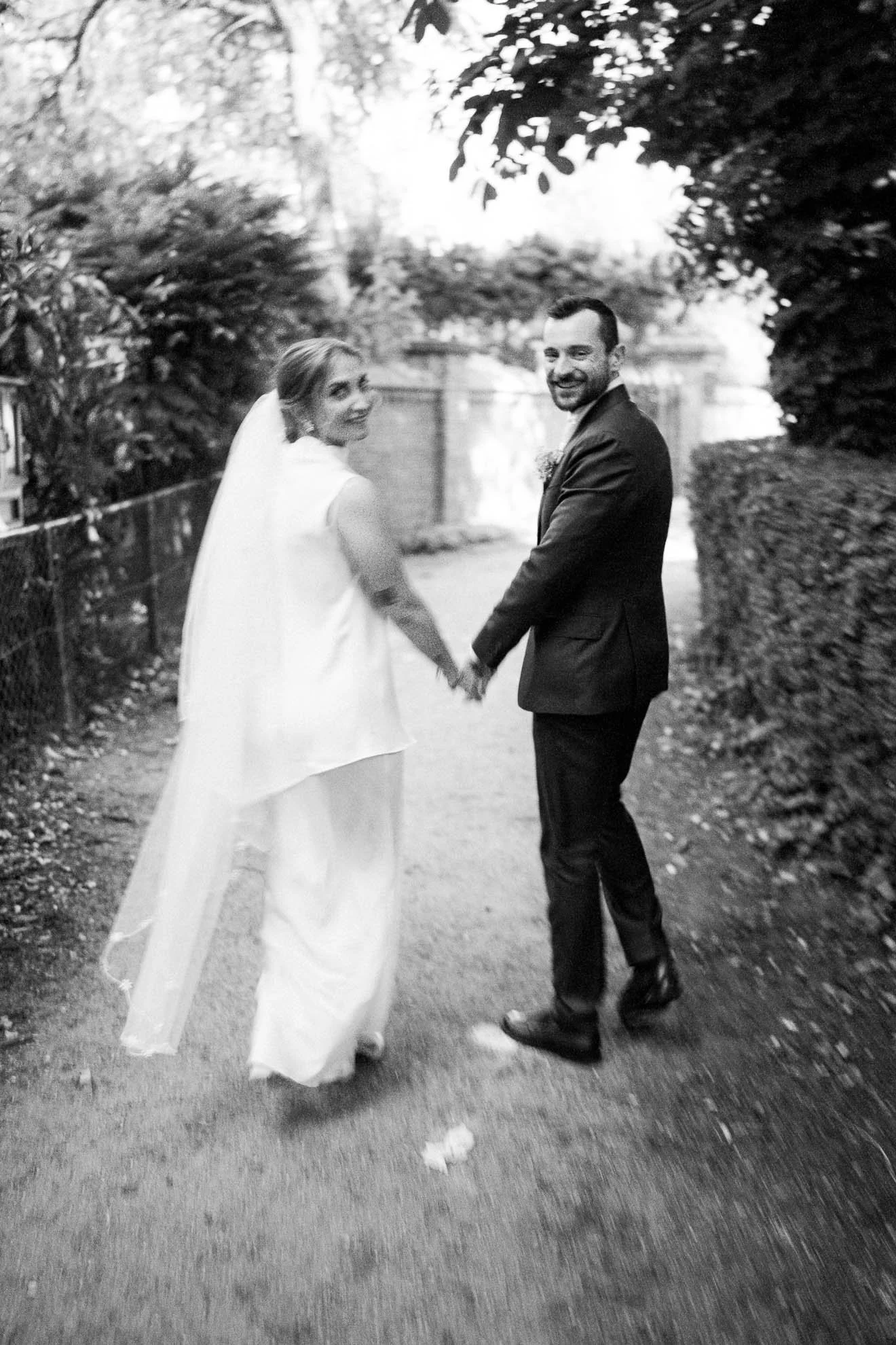A bride and groom holding hands and smiling at the camera on a pathway lined with trees and bushes, in outdoor wedding setting, black and white photograph.