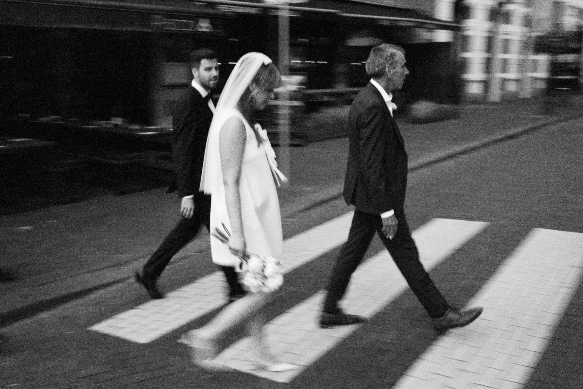 Black and white photo of three people crossing a street at a pedestrian crosswalk, including a bride in a wedding dress and veil, a man in a tuxedo, and another man in a suit. The photo has a motion blurred effect.