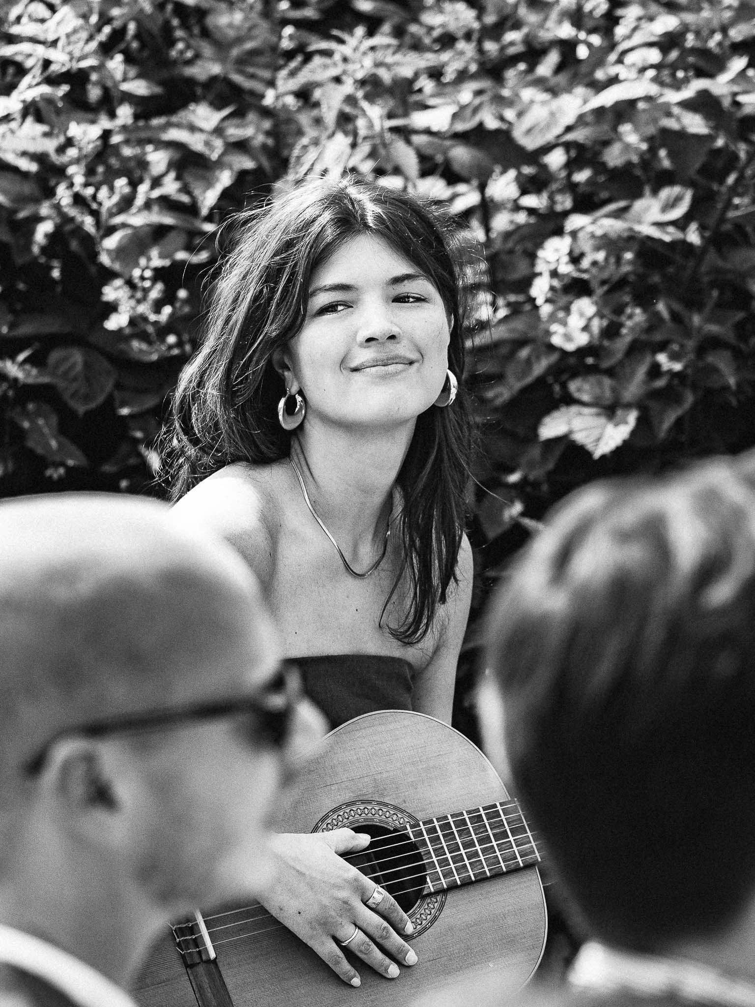 Zwarte en witte foto van een vrouw die gitaar speelt en glimlacht, met bladeren op de achtergrond, omringd door anderen.