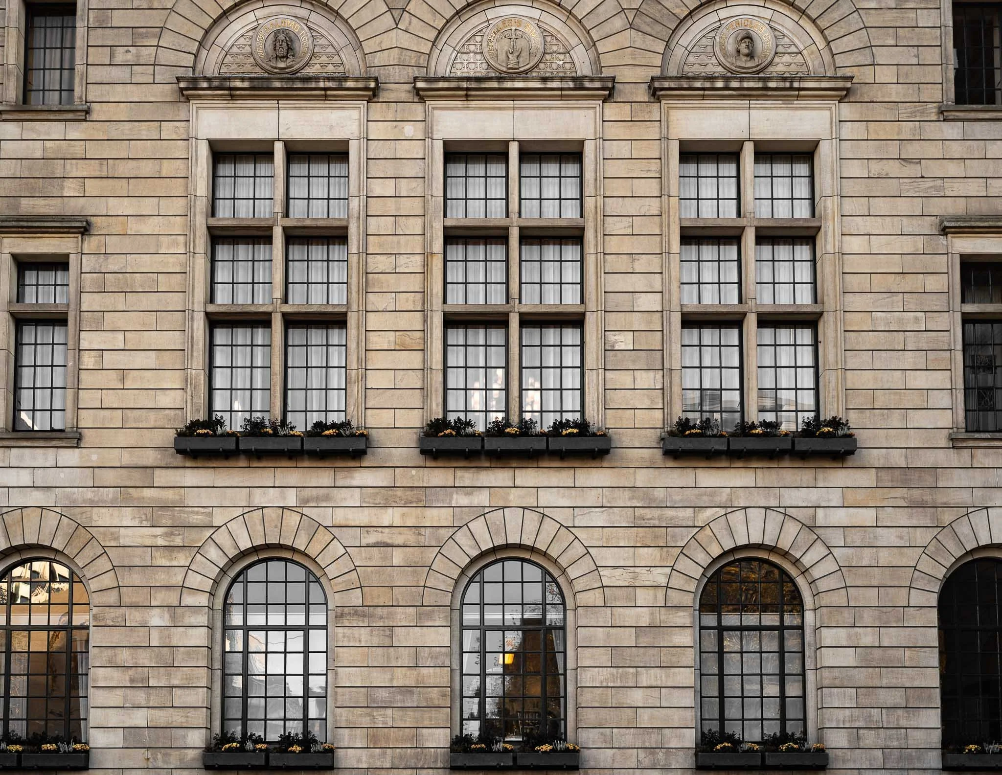 Gezicht op de gevel van een oud gebouw met grote ramen, bloemrijke vensterbanken en drie medailles of reliëfs boven de ramen.