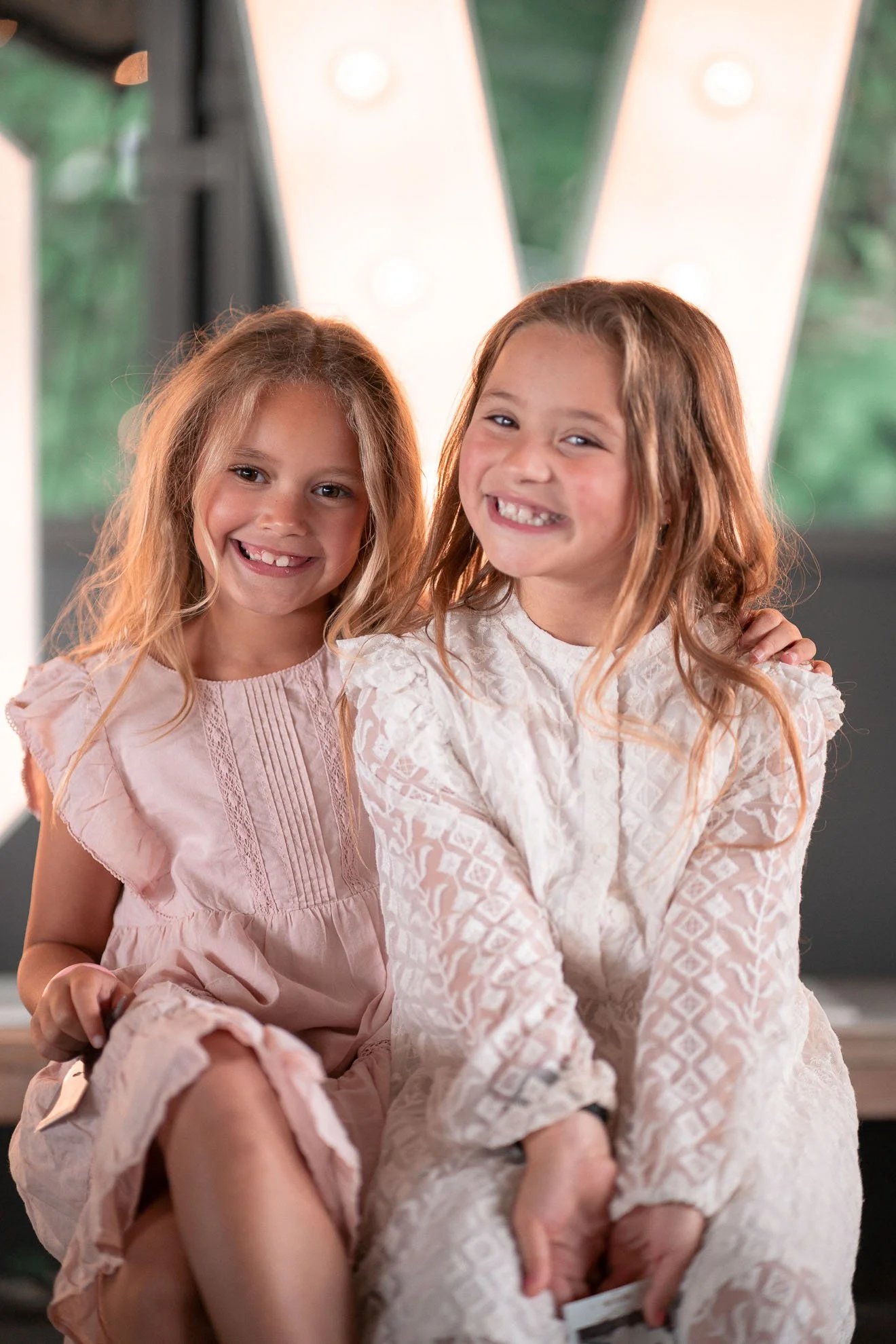 Two young girls with smiles, sitting closely together, both wearing light-colored dresses, with a large illuminated letter 'V' in the background.