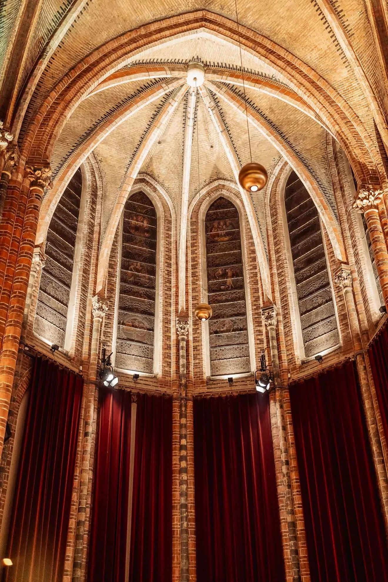 Interior of a Gothic-style church or cathedral with high arched brick ceiling, stained glass windows, and red velvet curtains.