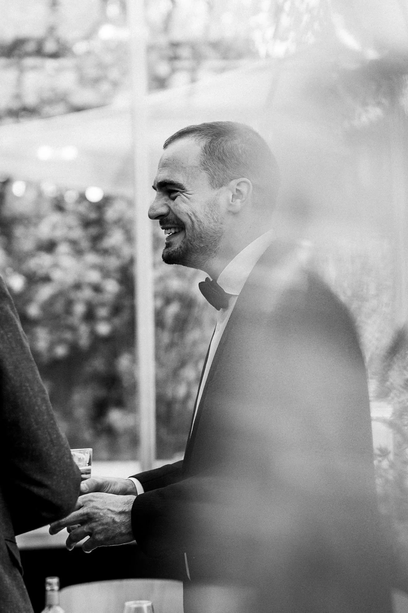 A smiling man in a tuxedo and bow tie holding a drink, engaging with someone else during a social event.