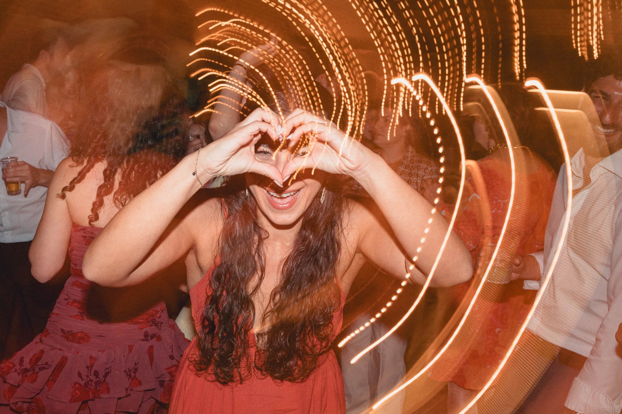 A woman with curly hair smiling and making a heart shape with her hands near her face at a party, surrounded by streaks of orange and yellow lights, with other people in the background enjoying the event.