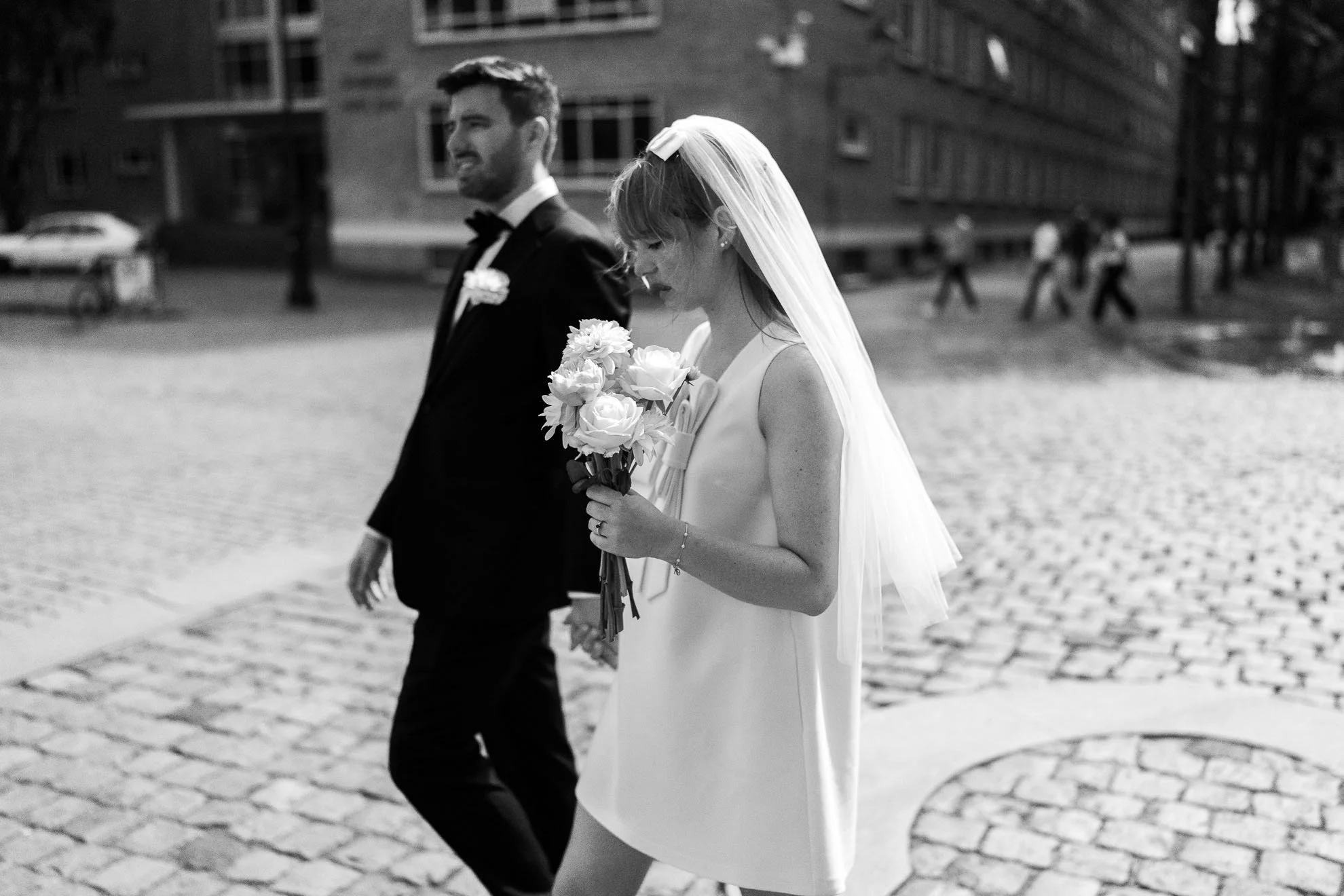 Black and white photo of a bride and groom walking together outdoors, the bride holding a bouquet of flowers, with both dressed in wedding attire.
