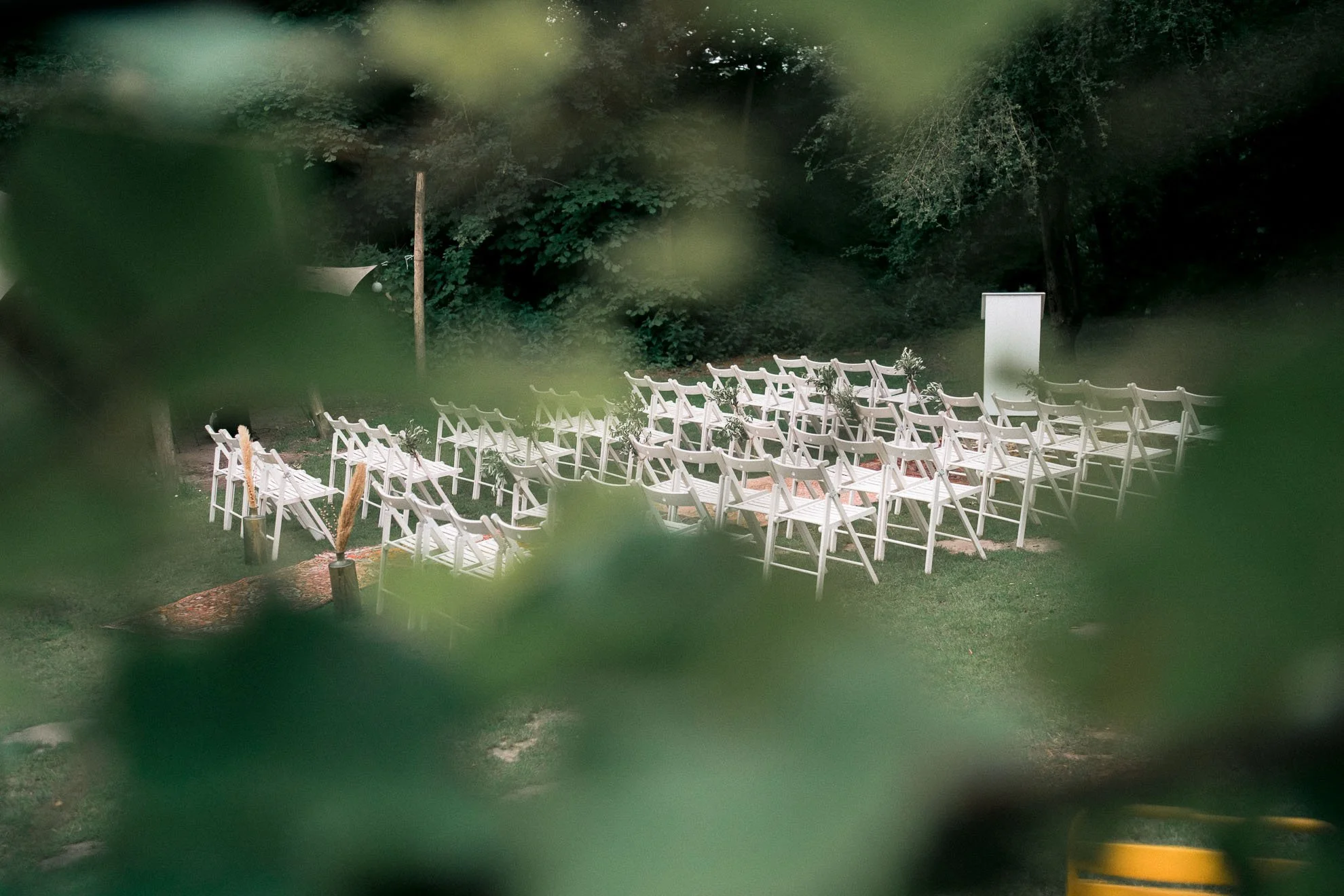 Outdoor setting with rows of white chairs arranged on grass, likely for a wedding or event, surrounded by trees and greenery.