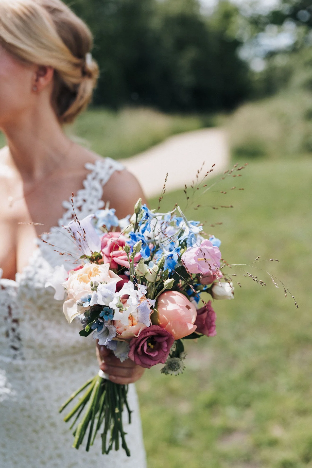 A woman in a white lace wedding dress holding a large bouquet of colorful flowers outdoors with green trees in the background.