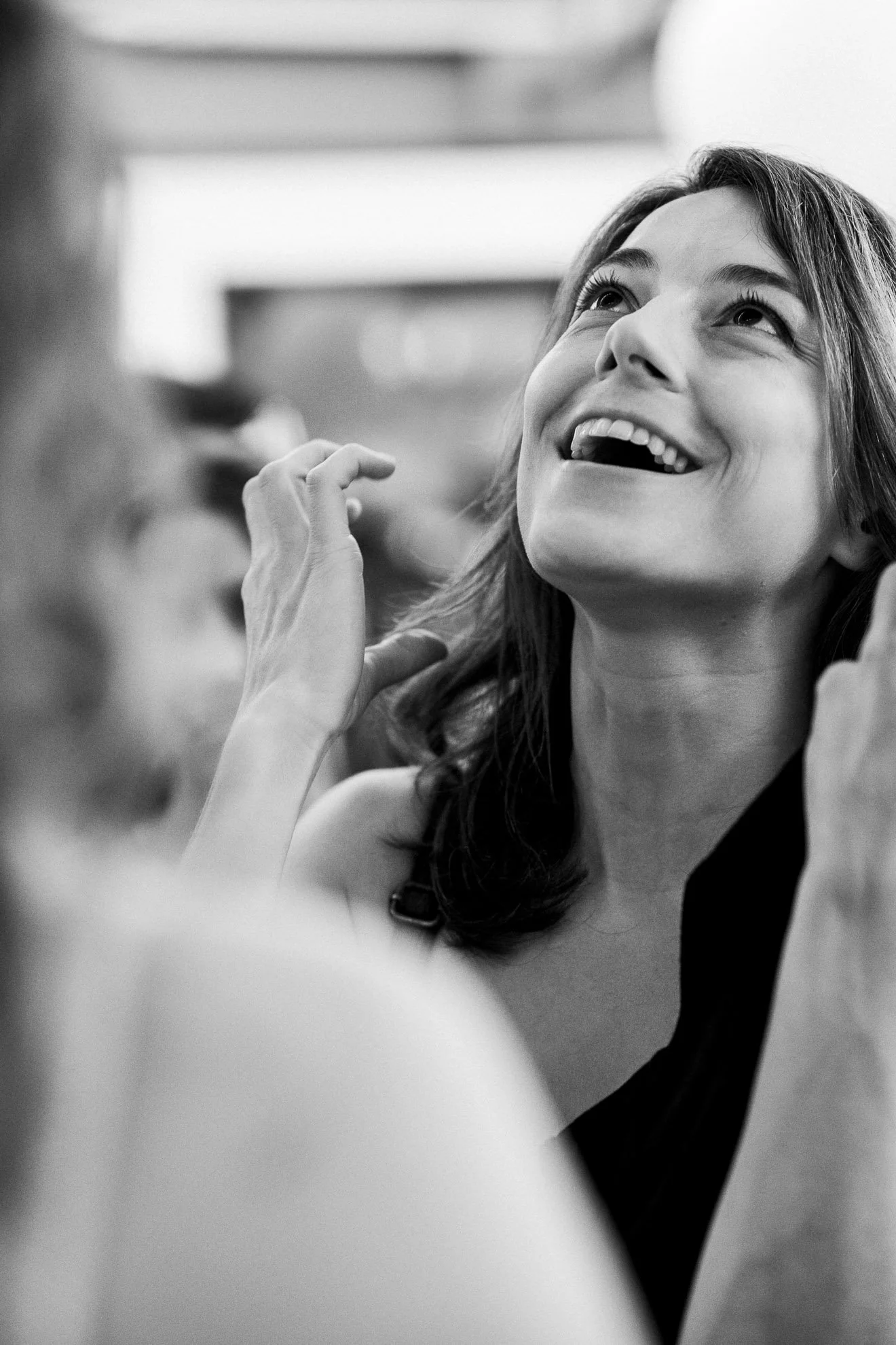 Black and white photo of a young woman smiling and looking upwards, with a hand near her face, and blurred person in the foreground.