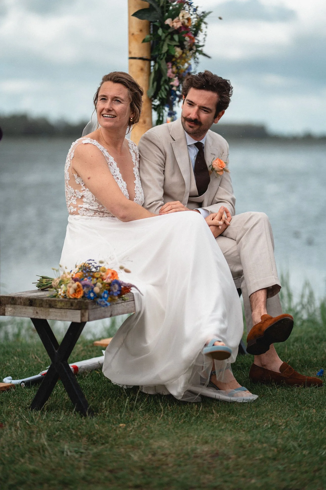 A bride and groom sitting together on a wooden bench outdoors during a lakeside wedding ceremony. The bride is in a white lace wedding dress with her veil, and the groom is in a light-colored suit with a boutonniere. They are holding hands and lookin