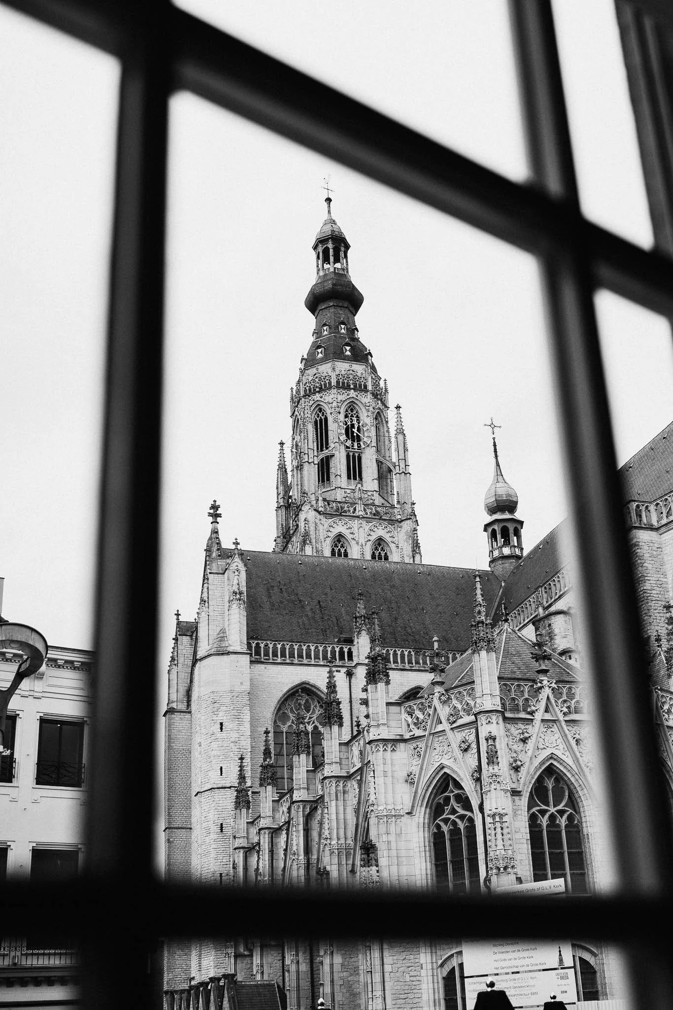 Zwarte en witte foto van een gotische kerk met hoge toren, gezien door een raam met rooster.
