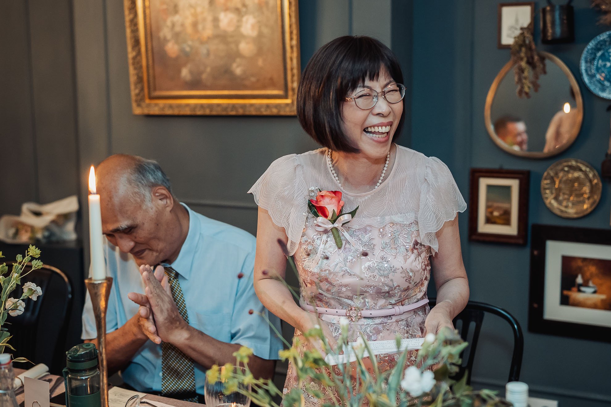 A woman with glasses, wearing a light pink dress with lace details, is smiling and standing at a table during a gathering. She has a corsage with a pink rose on her dress. There is a man with gray hair and glasses sitting next to her, with his eyes c