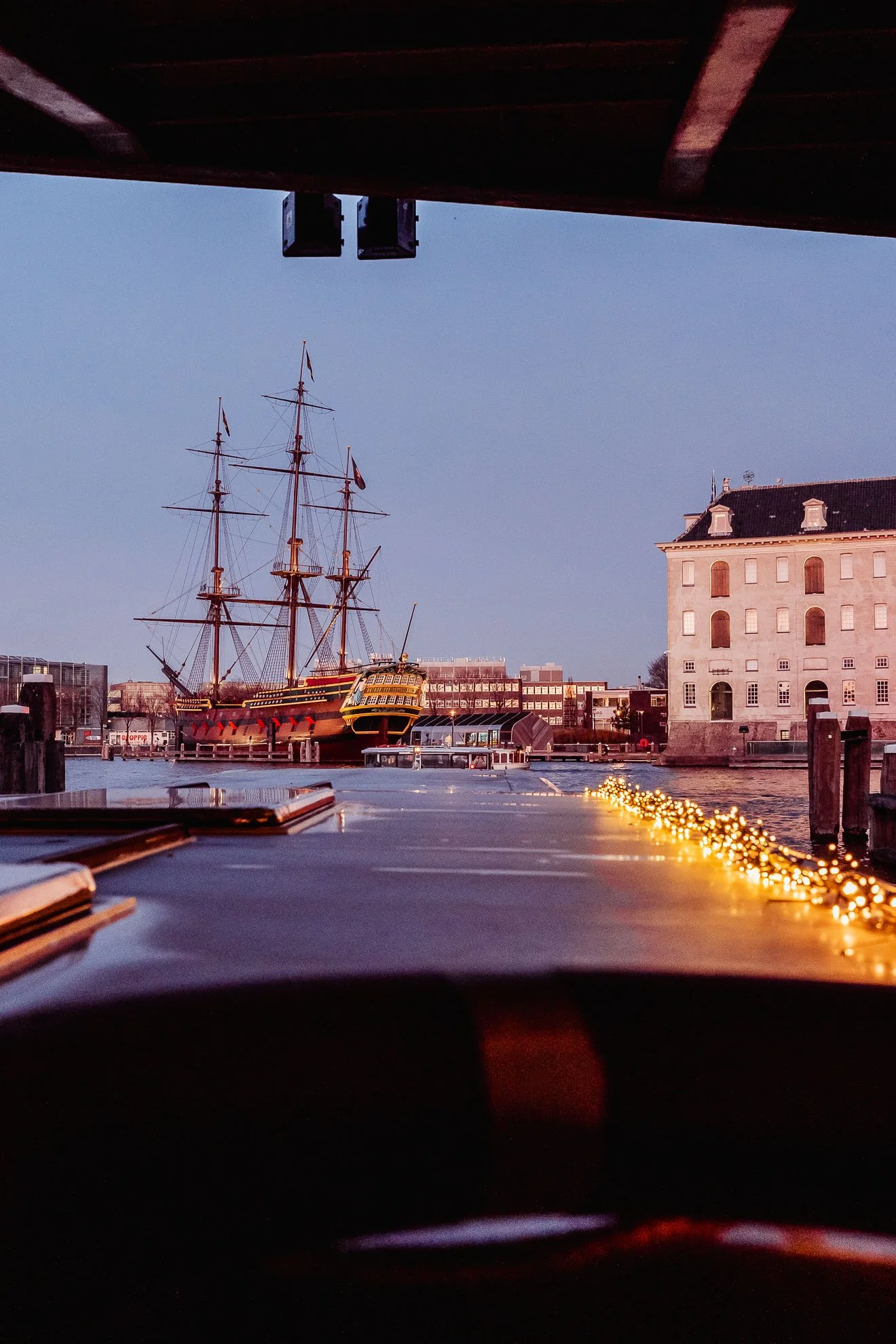 A view of a historical sailing ship docked at a harbor during twilight, with modern buildings and a large stone building in the background. The scene is viewed from underneath a wooden structure with string lights in the foreground.