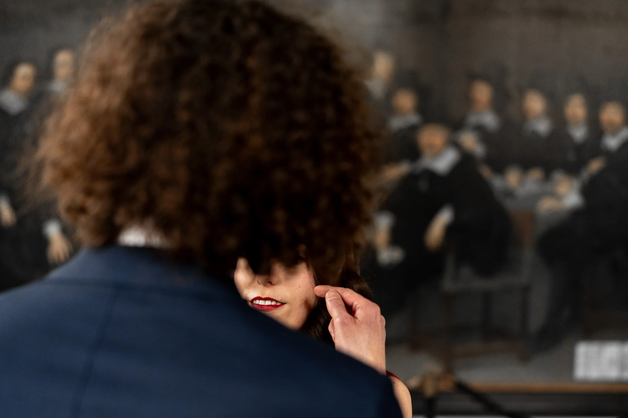 Een vrouw met rode lippen wordt door een persoon met kastanjebrouwe, krullende haar gekust, terwijl een portret van een groep mensen op de achtergrond hangt.