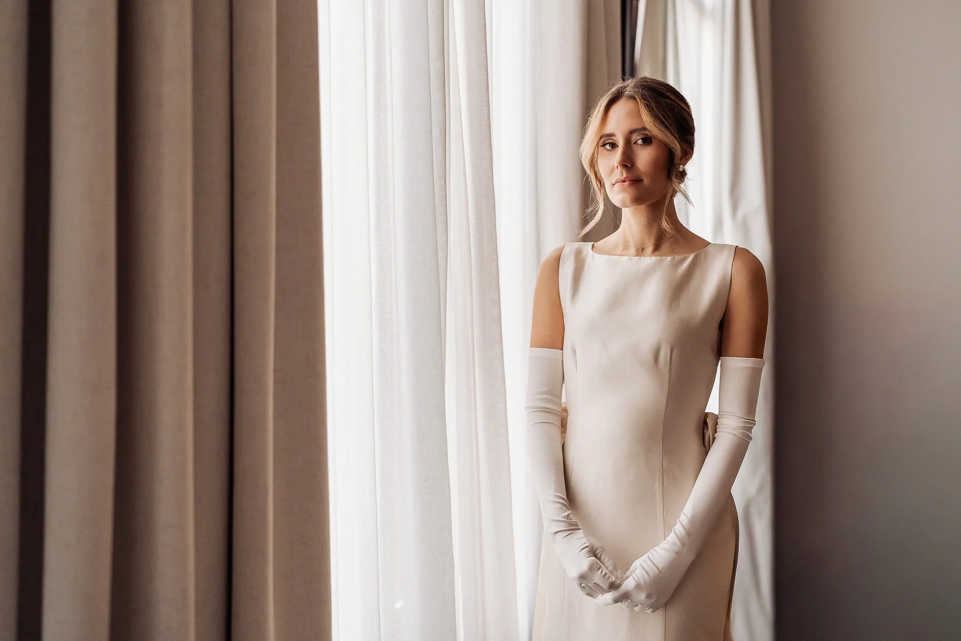 Vrouw in een witte trouwjurk en lange handschoenen staat bij een raam met beige gordijnen.