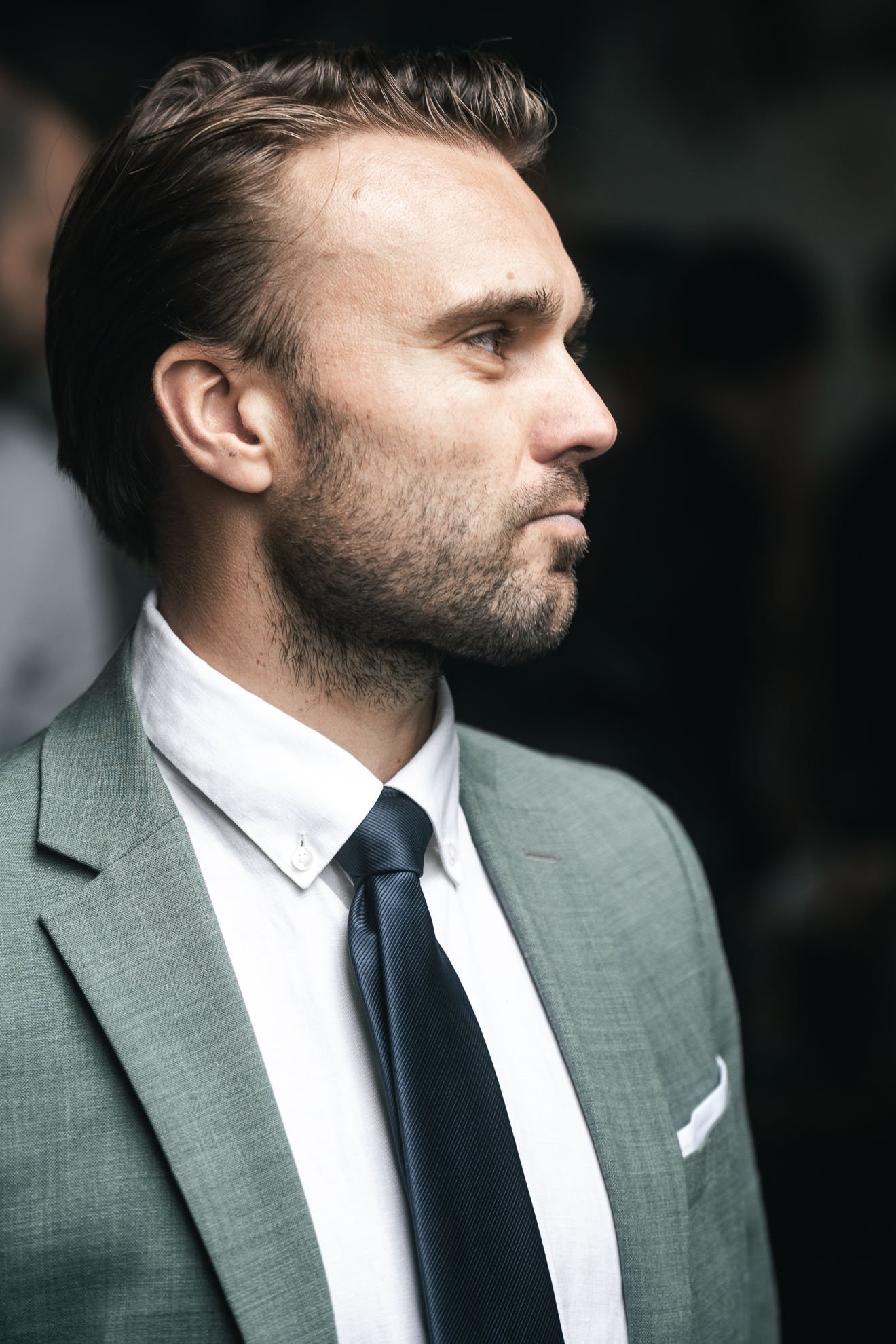 Side profile of a man in a gray suit, white shirt, and dark tie, with slicked-back hair and slight facial hair, standing against a dark background.