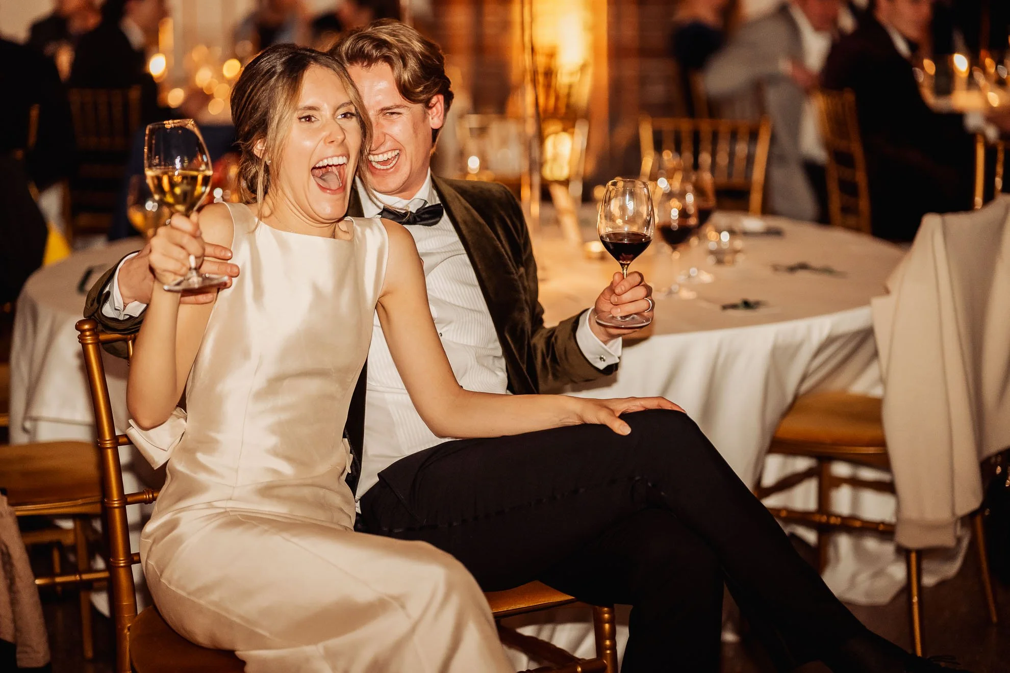 A man and woman celebrate at a formal event, laughing and holding glasses of wine, with a decorated banquet hall setting in the background.