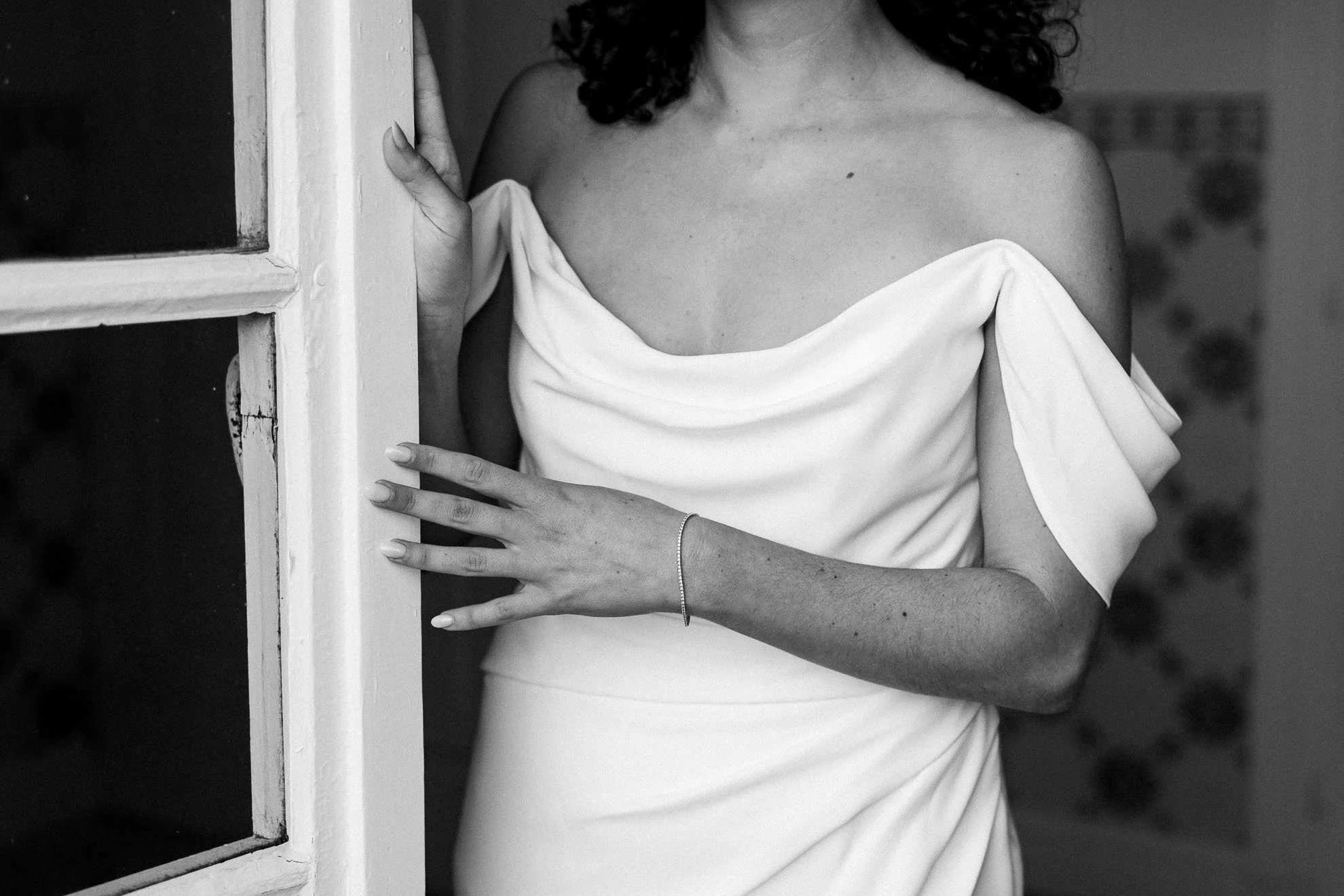 A woman wearing a strapless white dress is standing next to a window frame, her hand on the frame, with only her upper body visible in the black-and-white photo.