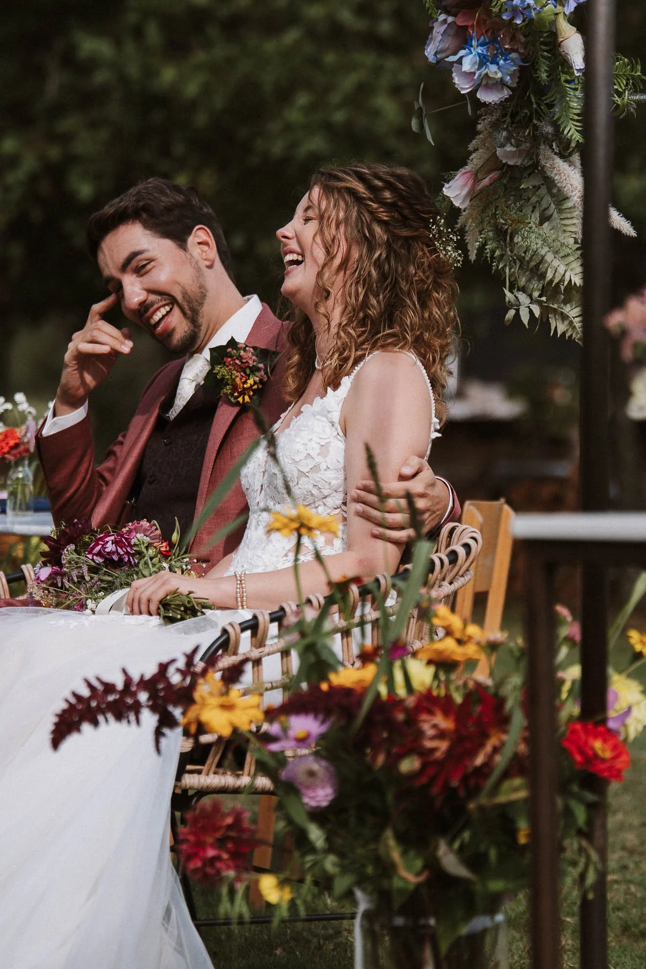 A joyful bride and groom sitting at a wedding reception outdoors, surrounded by floral decorations and greenery, smiling and laughing.