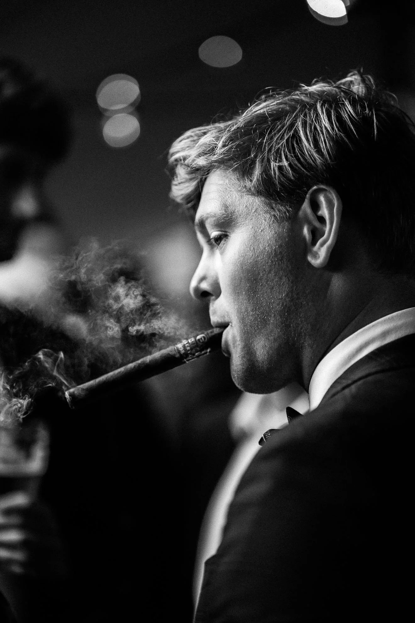 A black and white photo of a young man in formal attire smoking a cigar with smoke swirling around.