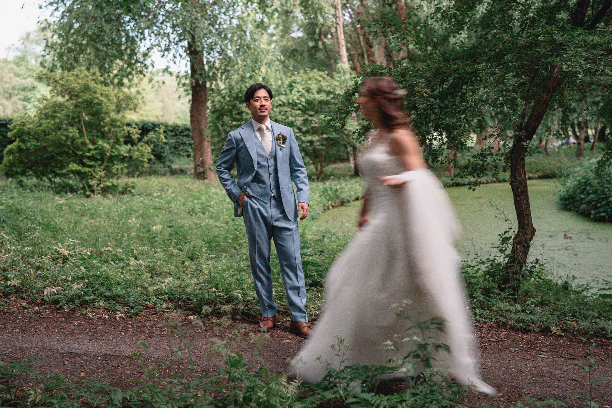 A man in a blue suit standing in a park or garden surrounded by trees, with a woman in a white dress blurred in motion in the foreground.