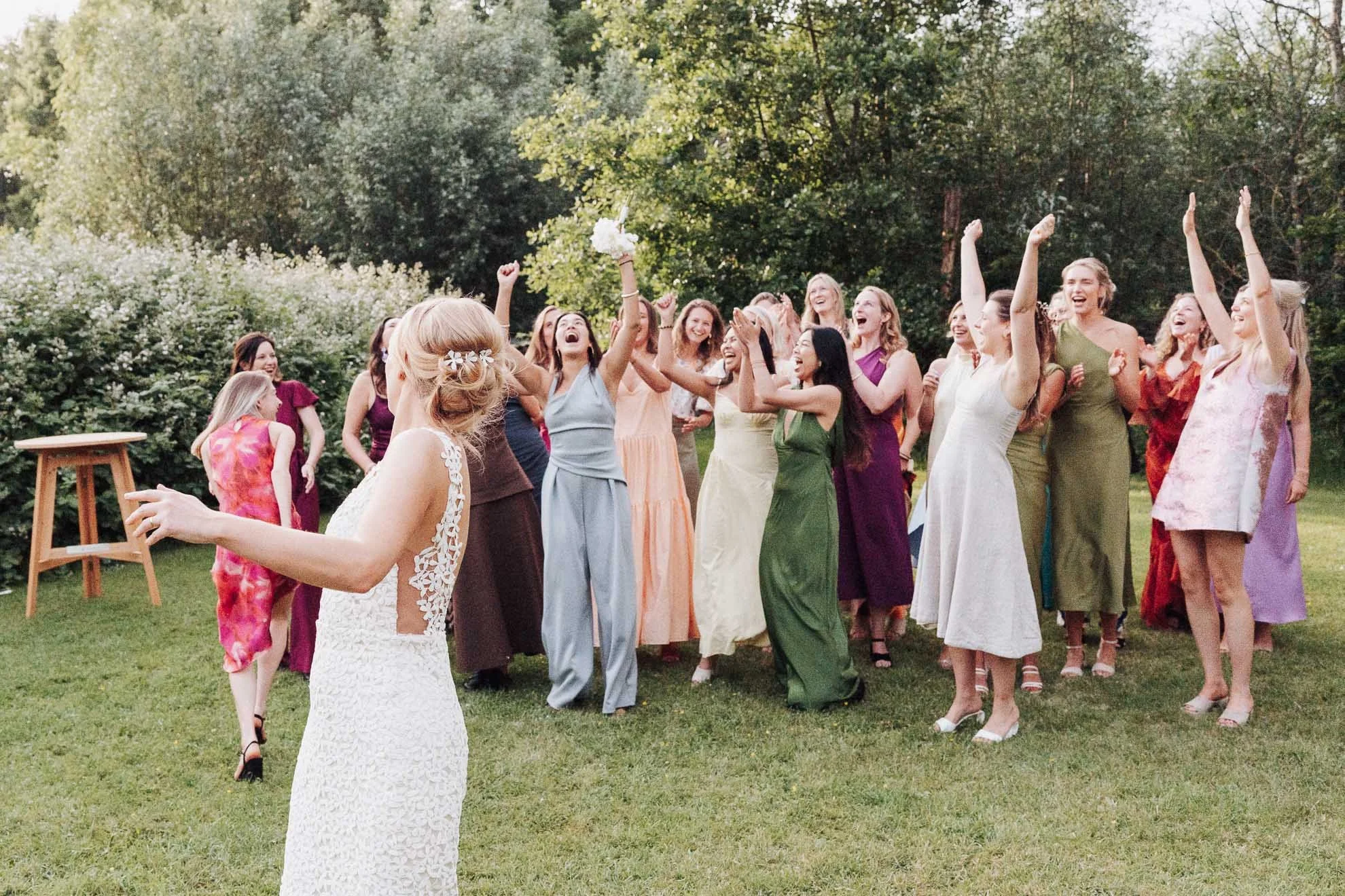A wedding celebration outdoors with women dressed in colorful evening gowns, dancing and cheering on a sunny day, surrounded by trees and greenery.