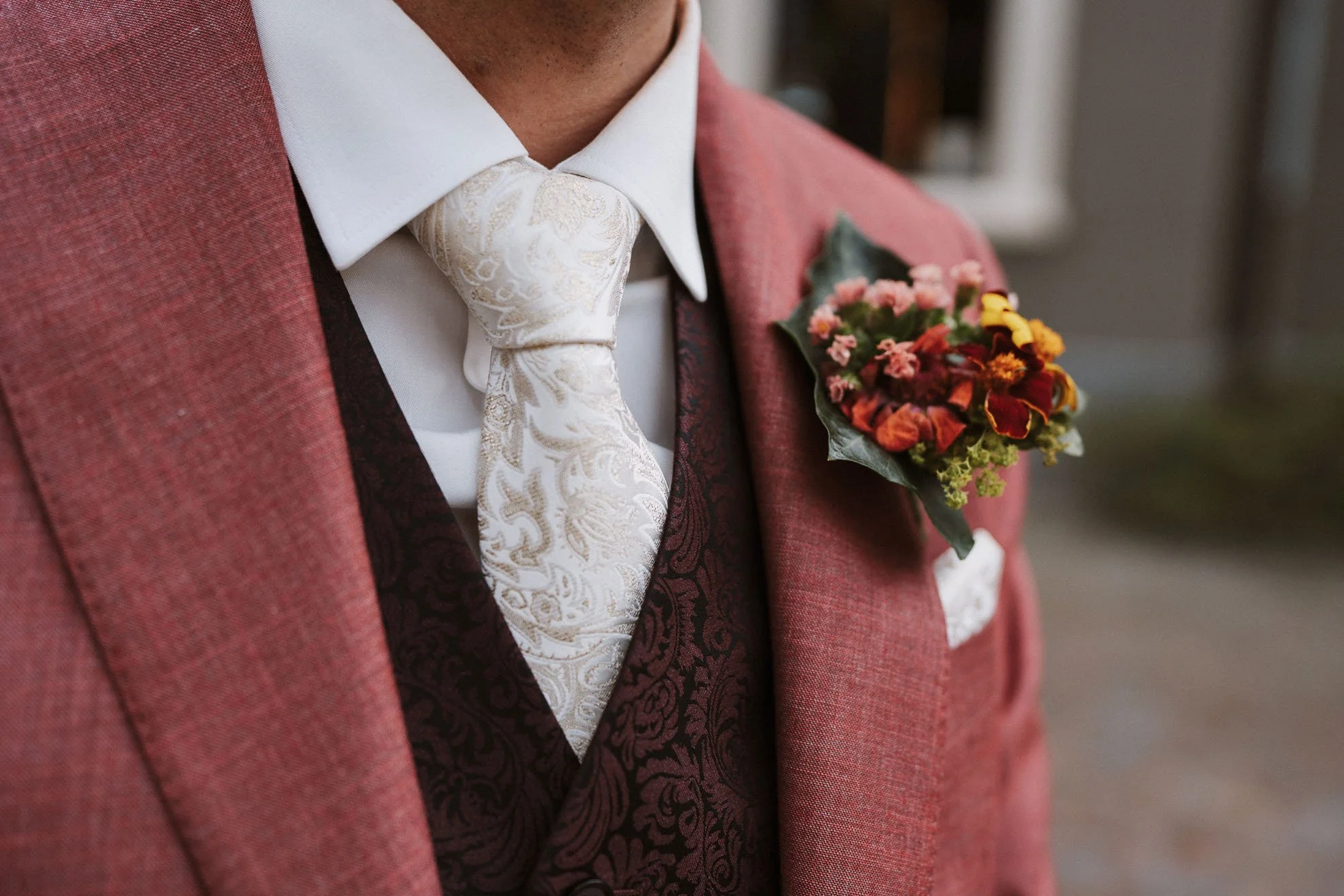 Close-up of a man in a red suit, white shirt, ornate cream-colored tie, and floral boutonniere on his lapel.