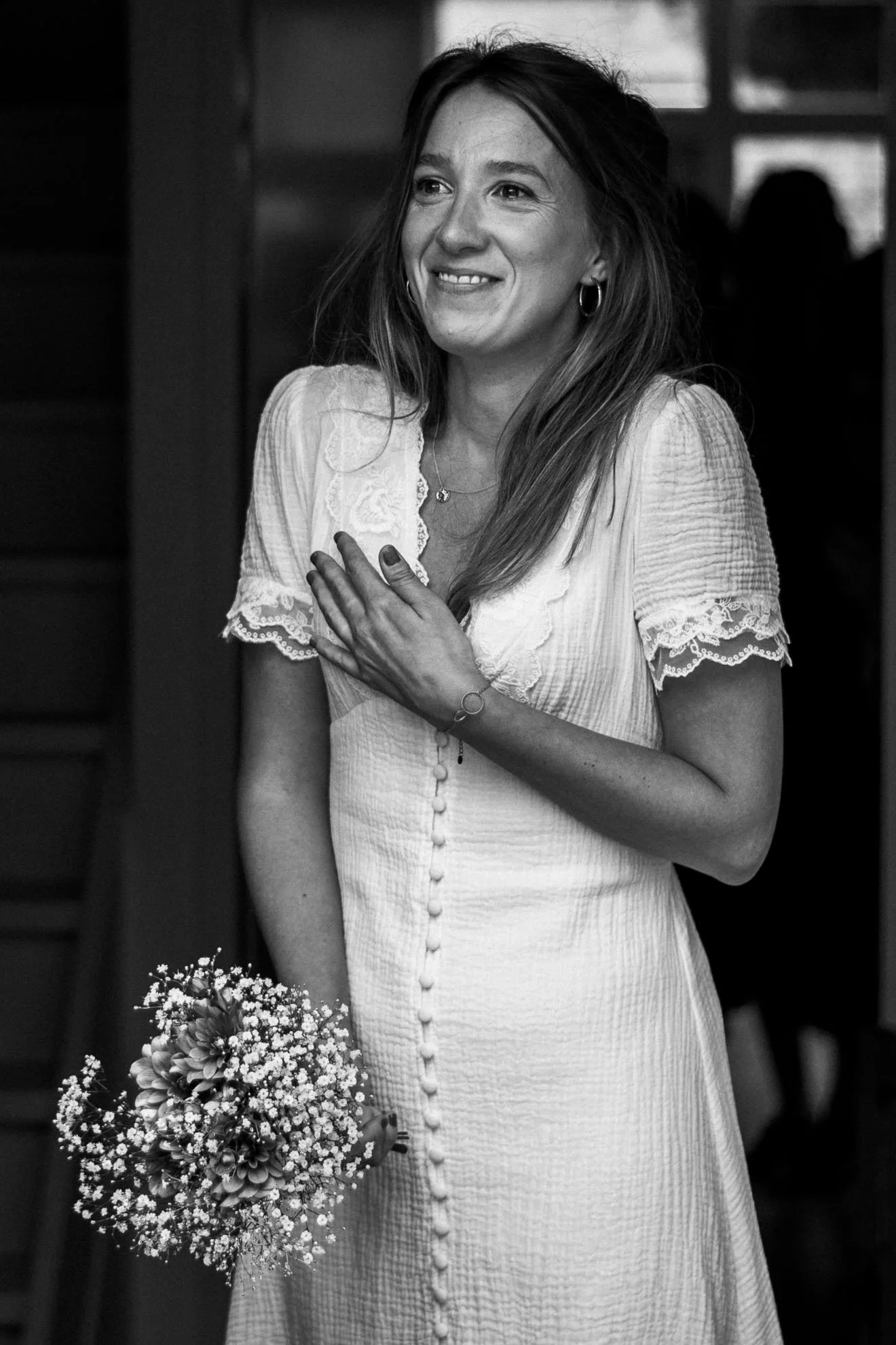 A woman in a white dress holding a bouquet of flowers, smiling, in a dark room with windows in the background.