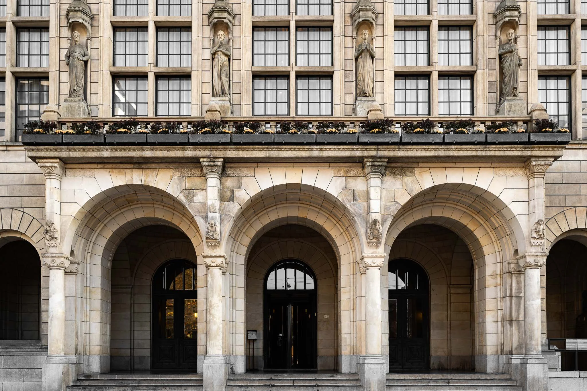 De façade van een gebouw met grote bogen en drie ingangen, versierd met beelden en bloemen op het balkon.