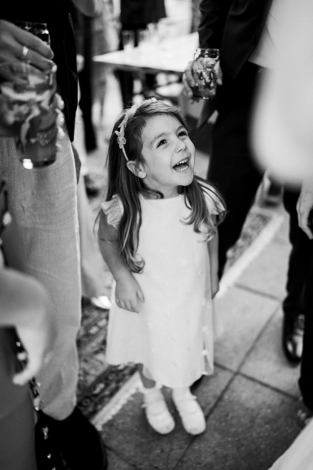 Een jonge meisje met lang haar en een diadeem, lachend en kijkend naar iemand die haar boven haar hoofd houdt, in een drukke omgeving met mensen en tafels.