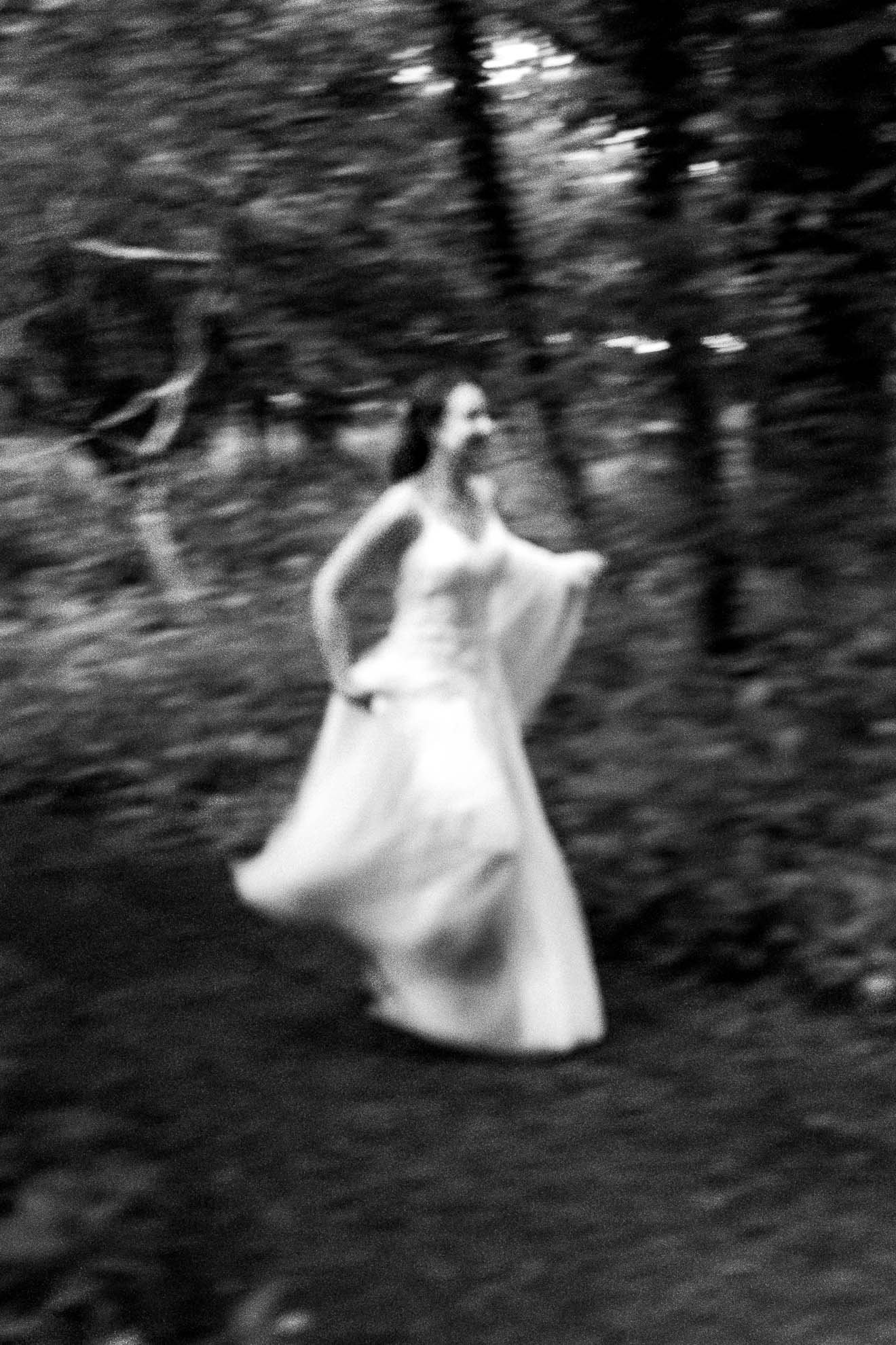A black and white photo of a woman in a flowing gown walking through a forest with blurred trees in the background.