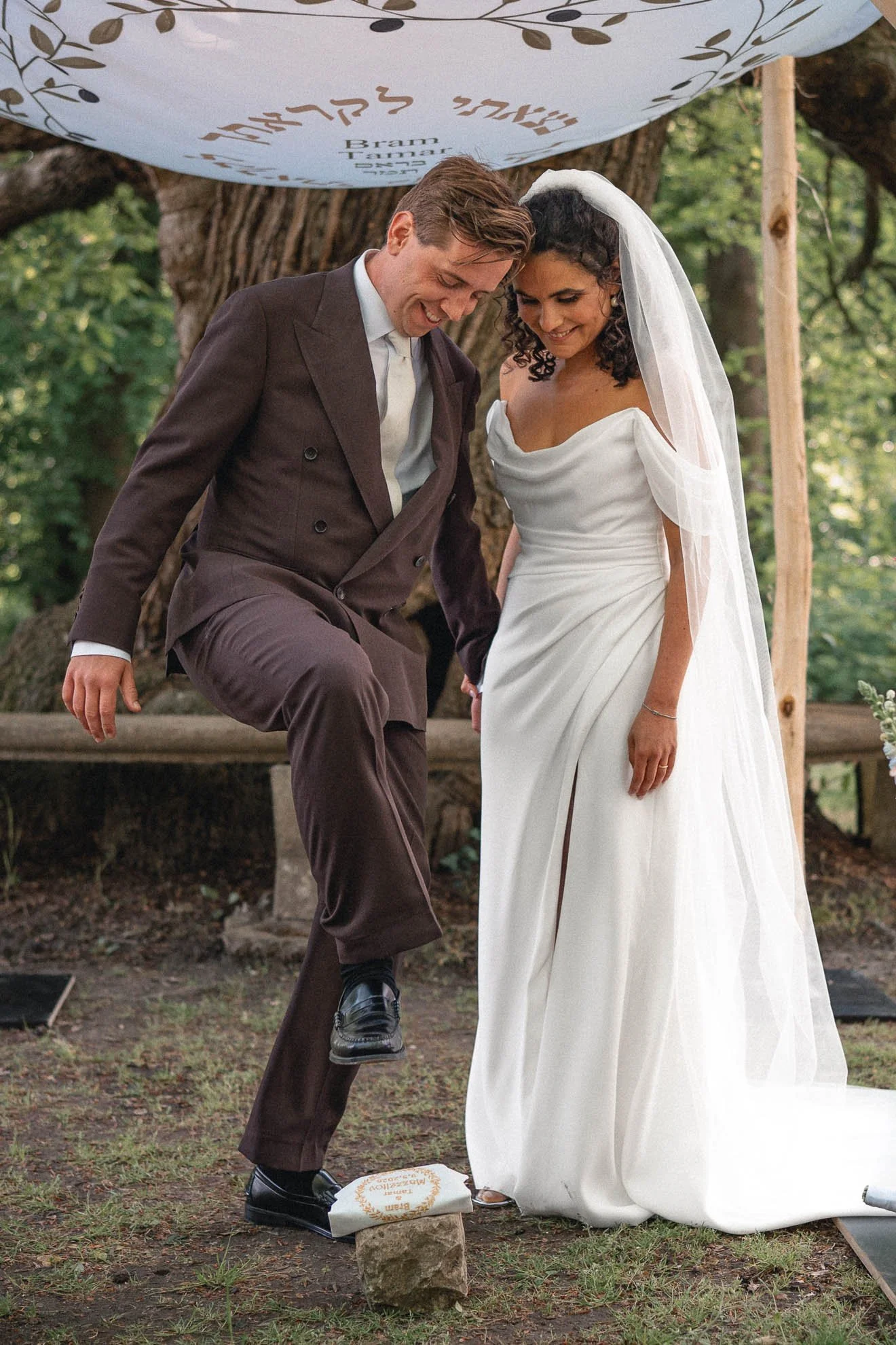 A bride and groom smiling during their outdoor wedding ceremony, with the groom stepping on a stone on a paper walk path.