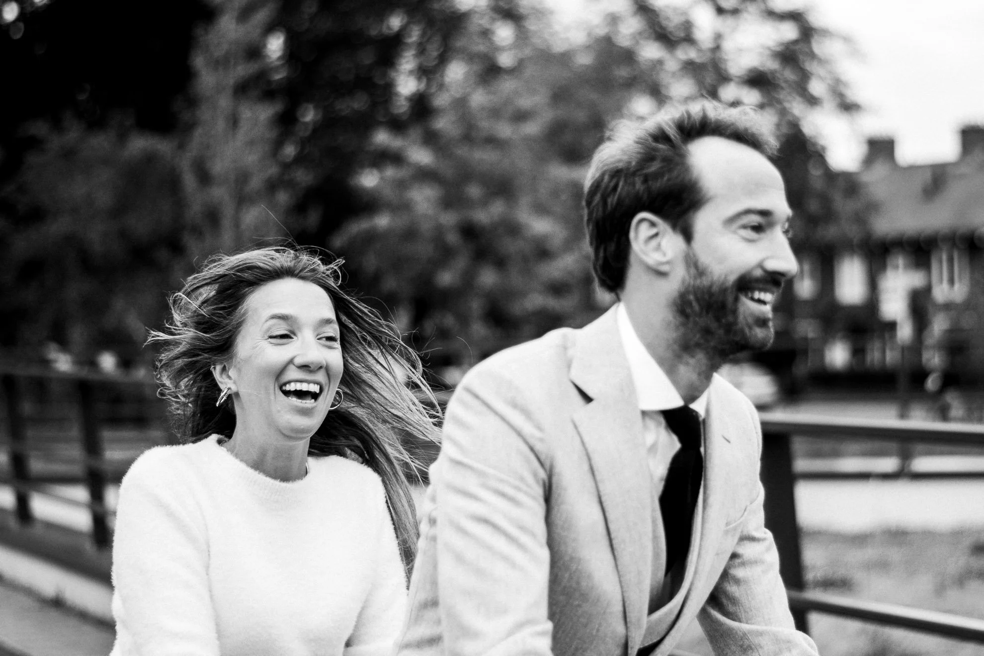 A black-and-white photo of a happy woman and man riding a bicycle outside. The woman has long hair and is smiling, while the man has a beard and is also smiling. They are enjoying a joyful moment together.