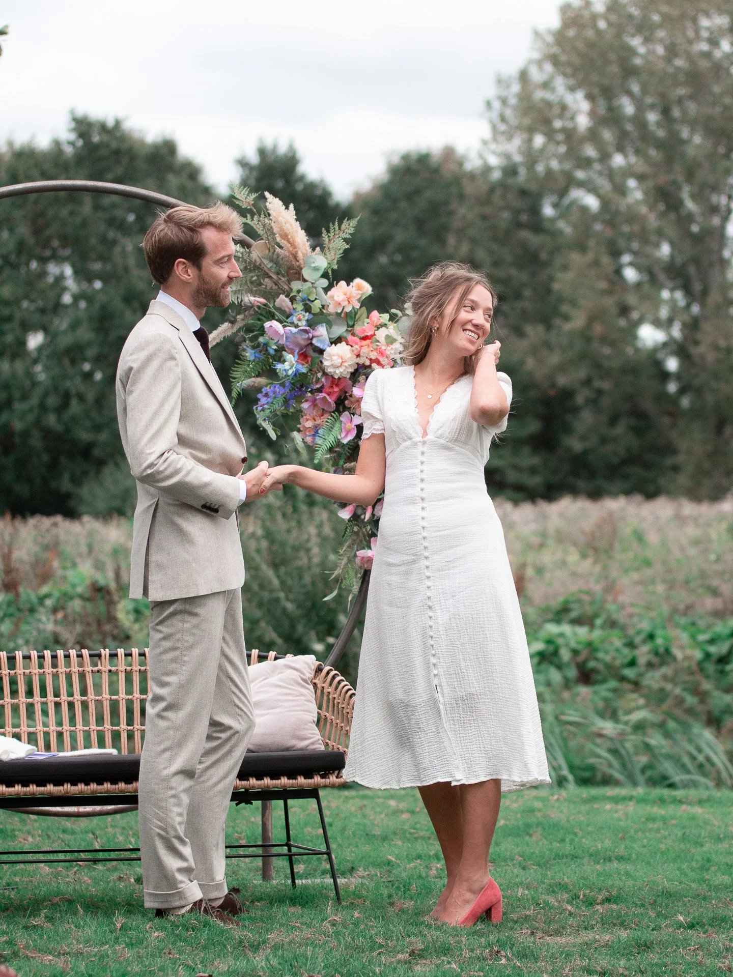 Een stel op haar bruiloft, buiten, met bloemen en een tuin in de achtergrond, ze lachen en houden elkaars hand vast.