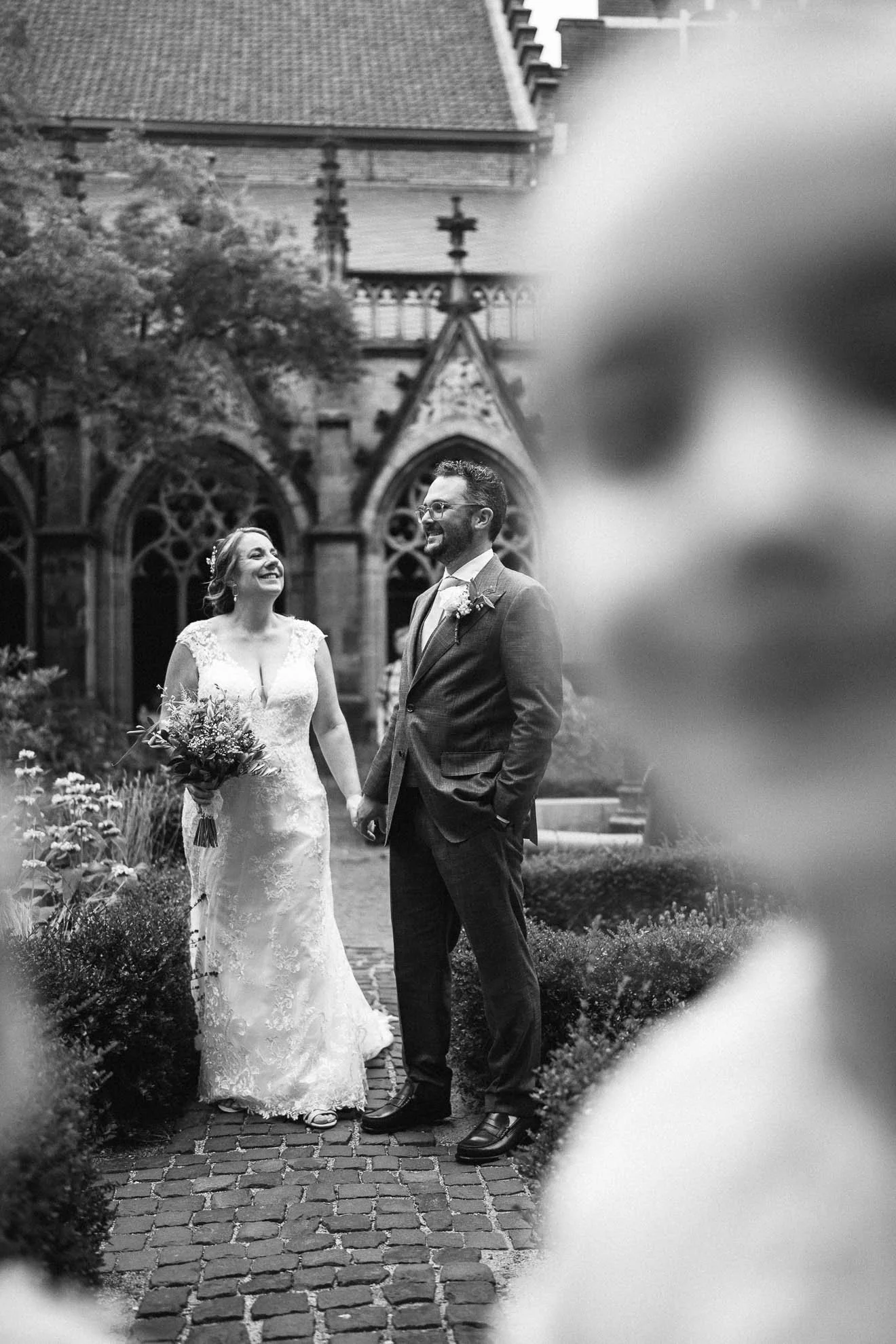 Black and white photo of a bride and groom holding hands and smiling at each other outside a church. The bride is wearing a lace wedding dress and holding a bouquet; the groom is dressed in a suit with glasses and a boutonniere. The church has gothic