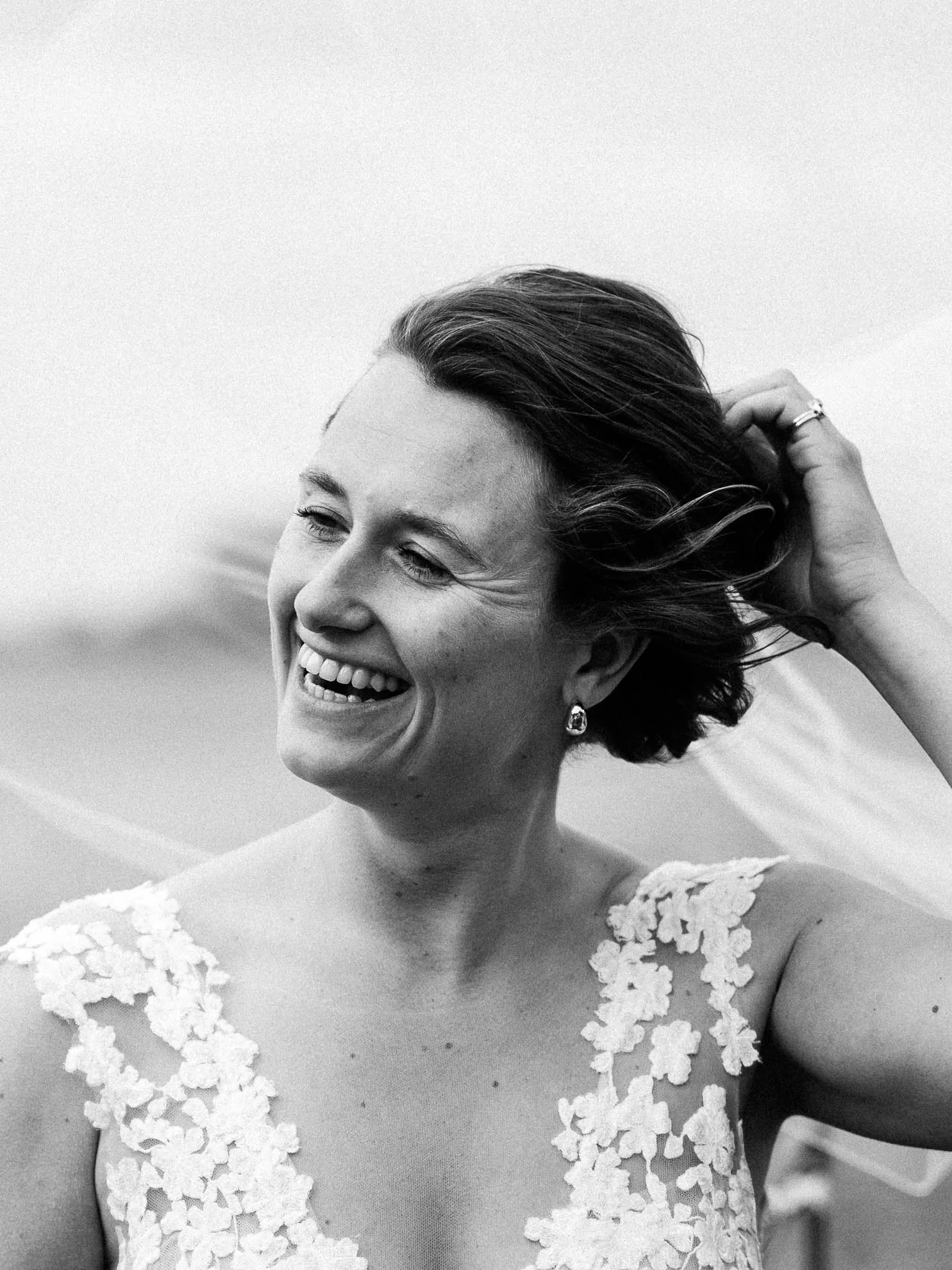 Black and white photo of a woman smiling, wearing a dress with lace details on the shoulders, her hair styled in waves, and holding her hair back with one hand.
