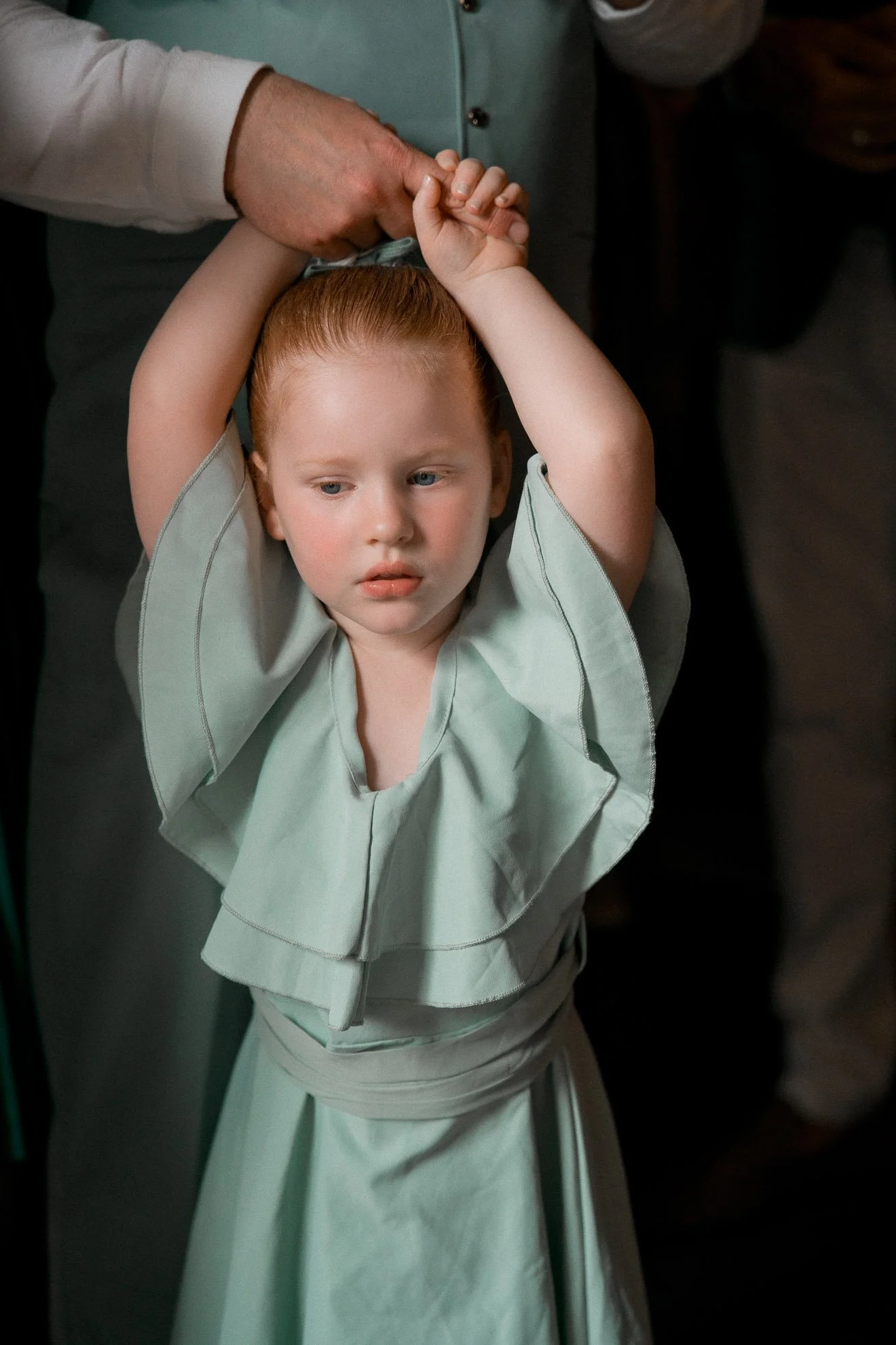 A young child with red hair and light skin standing on a toilet with the lid down, holding their hands above their head, and an adult hand gently holding the child's hand.