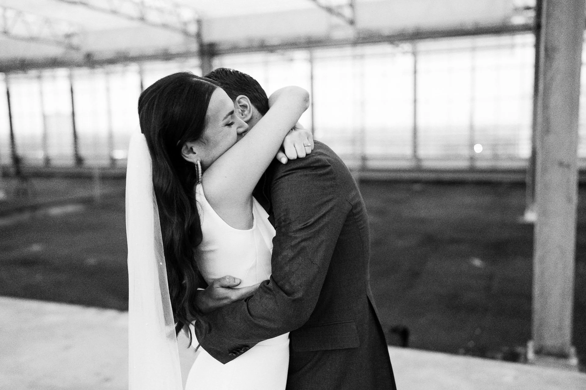 Een zwart-wit foto van een bruid en bruidegom die elkaar innig omhelzen en kussen in een lichte, open ruimte met grote ramen.