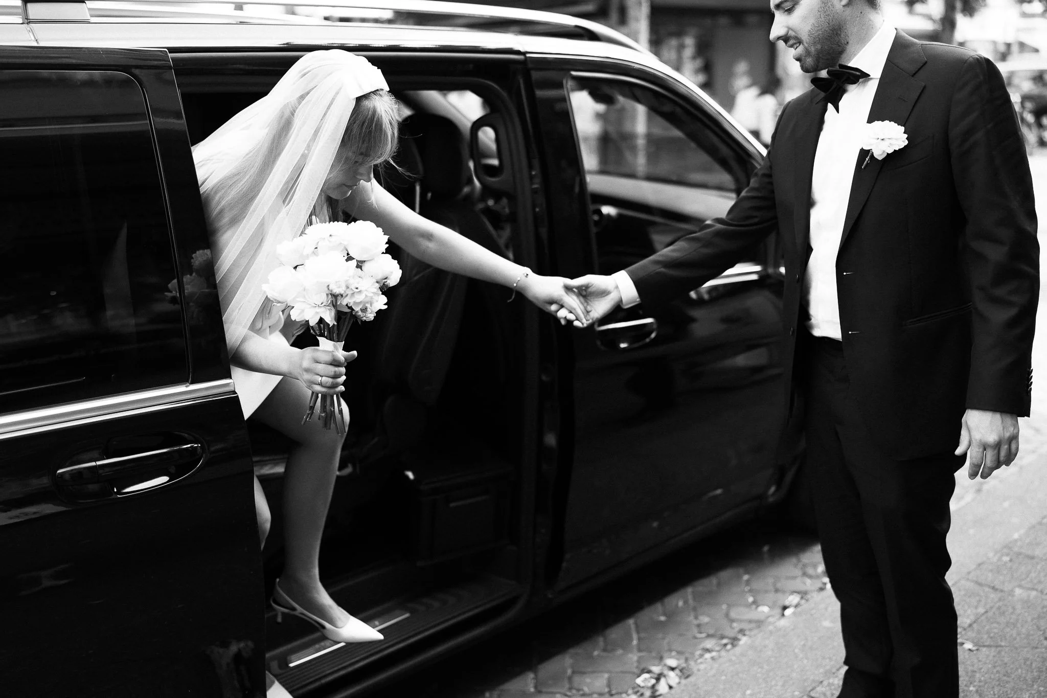 Bruidegom helpt bruid uit de auto terwijl hij haar hand aanneemt, op straat in zwart-wit.
