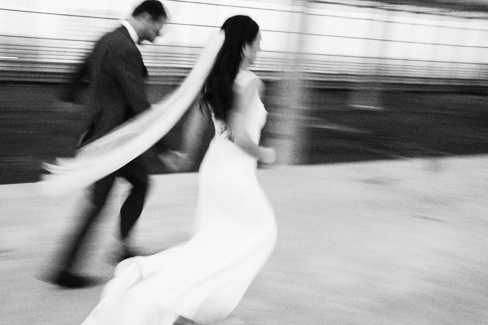 A black-and-white photograph of a man in a suit and a woman in a wedding dress walking in an urban setting with horizontal lines in the background. The couple appears to be holding hands and walking away from the camera.
