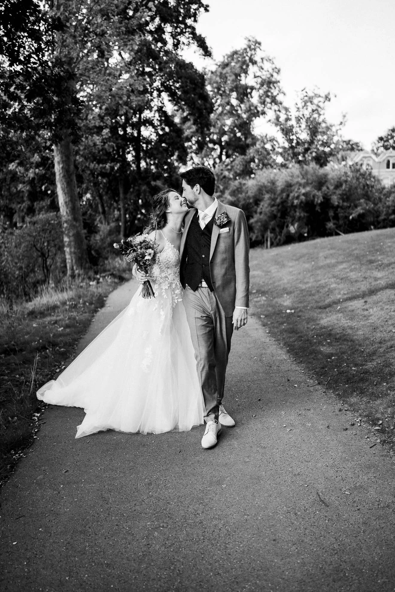 Black and white photo of a bride and groom walking together outside, smiling and looking at each other. The bride holds a bouquet, and the groom wears a suit with a boutonniere. Trees and a building are visible in the background.