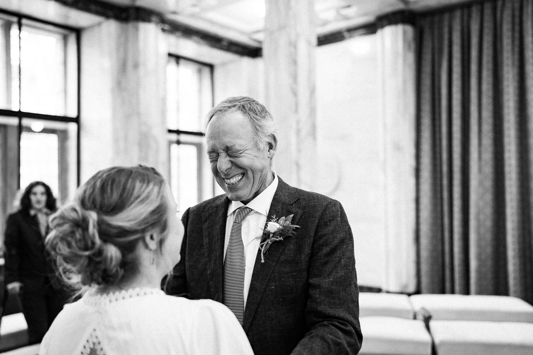 Een man en een vrouw lachen en kijken elkaar aan tijdens een ceremonie in een grote kamer met grote ramen en gordijnen.