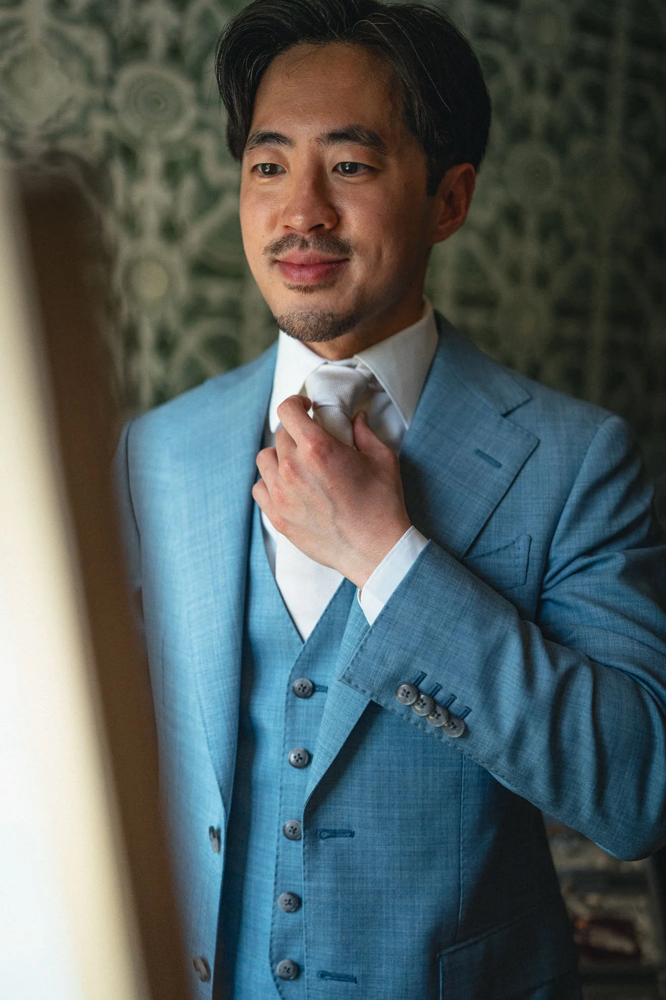 A man adjusting his tie, dressed in a light blue suit, white shirt, and standing next to a mirror with a patterned wall in the background.