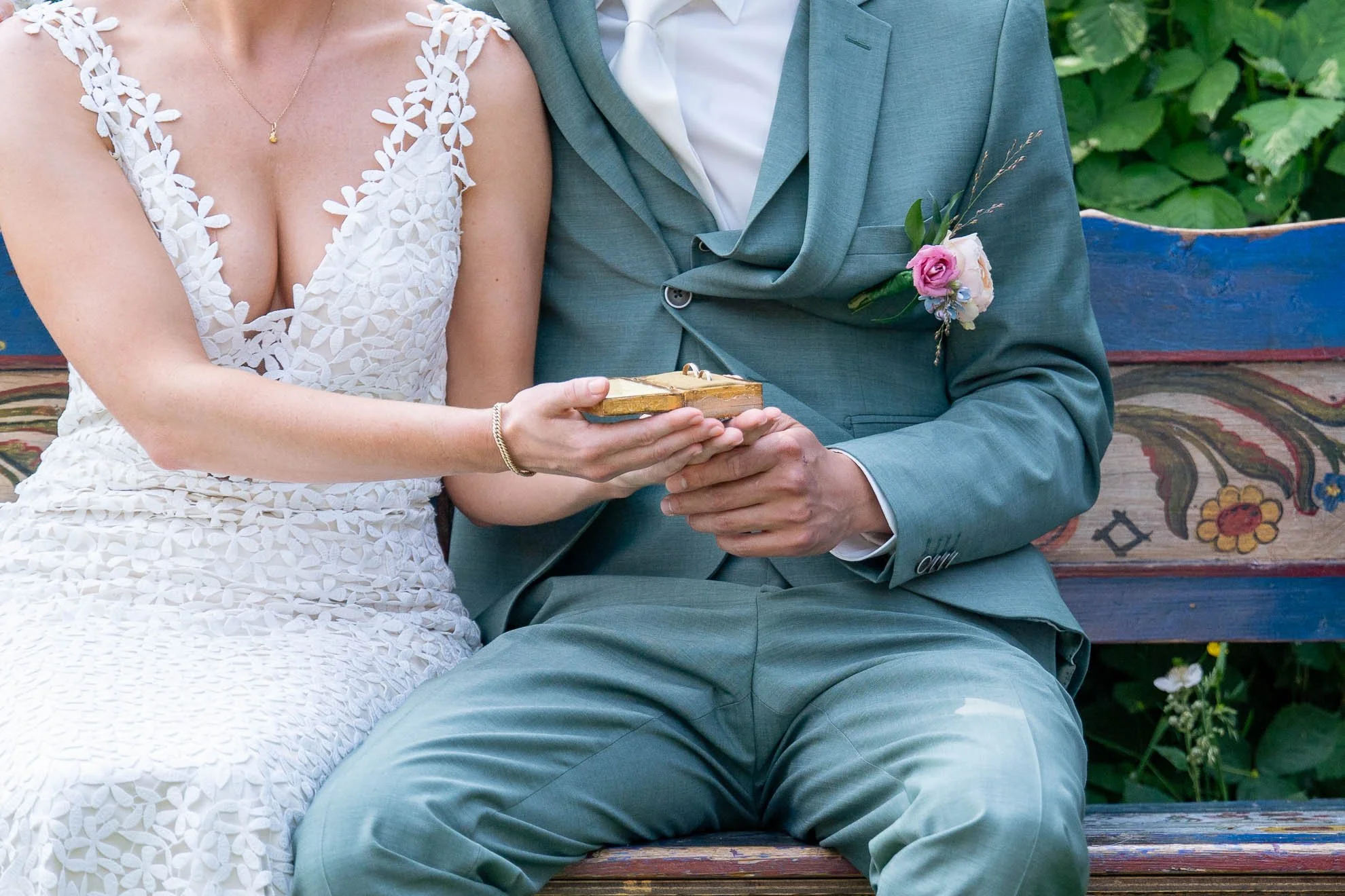 A bride and groom sitting on a wooden bench, holding a small wooden box, surrounded by green foliage.