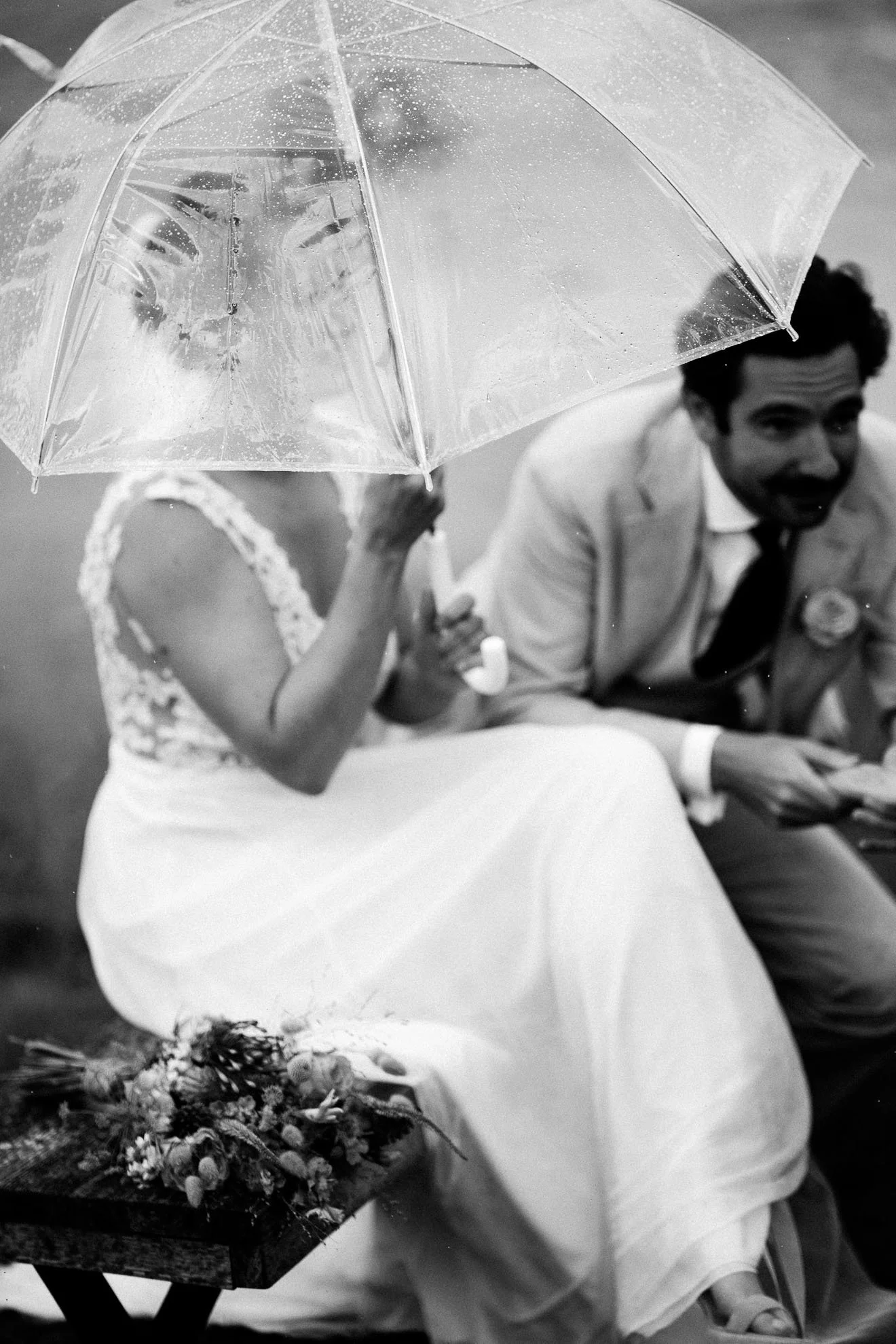 A woman in a white dress with lace sleeves and gloves sitting next to a man in a suit and tie, holding an umbrella decorated with rain droplets. In front, there is a wooden table with a large bouquet of flowers.