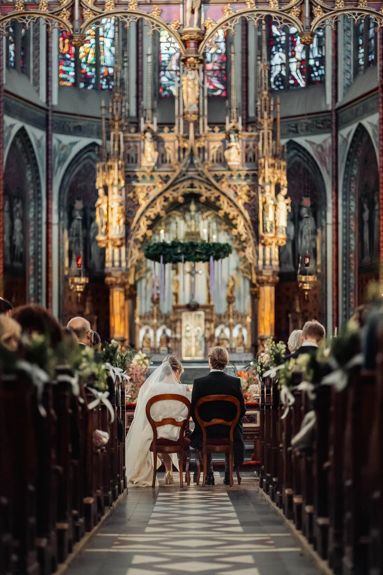 A bride and groom sit facing the altar in a decorated church for their wedding ceremony. The church features intricate stained glass windows, gothic architecture, and ornate gold detailing. Guests are seated along the pews.