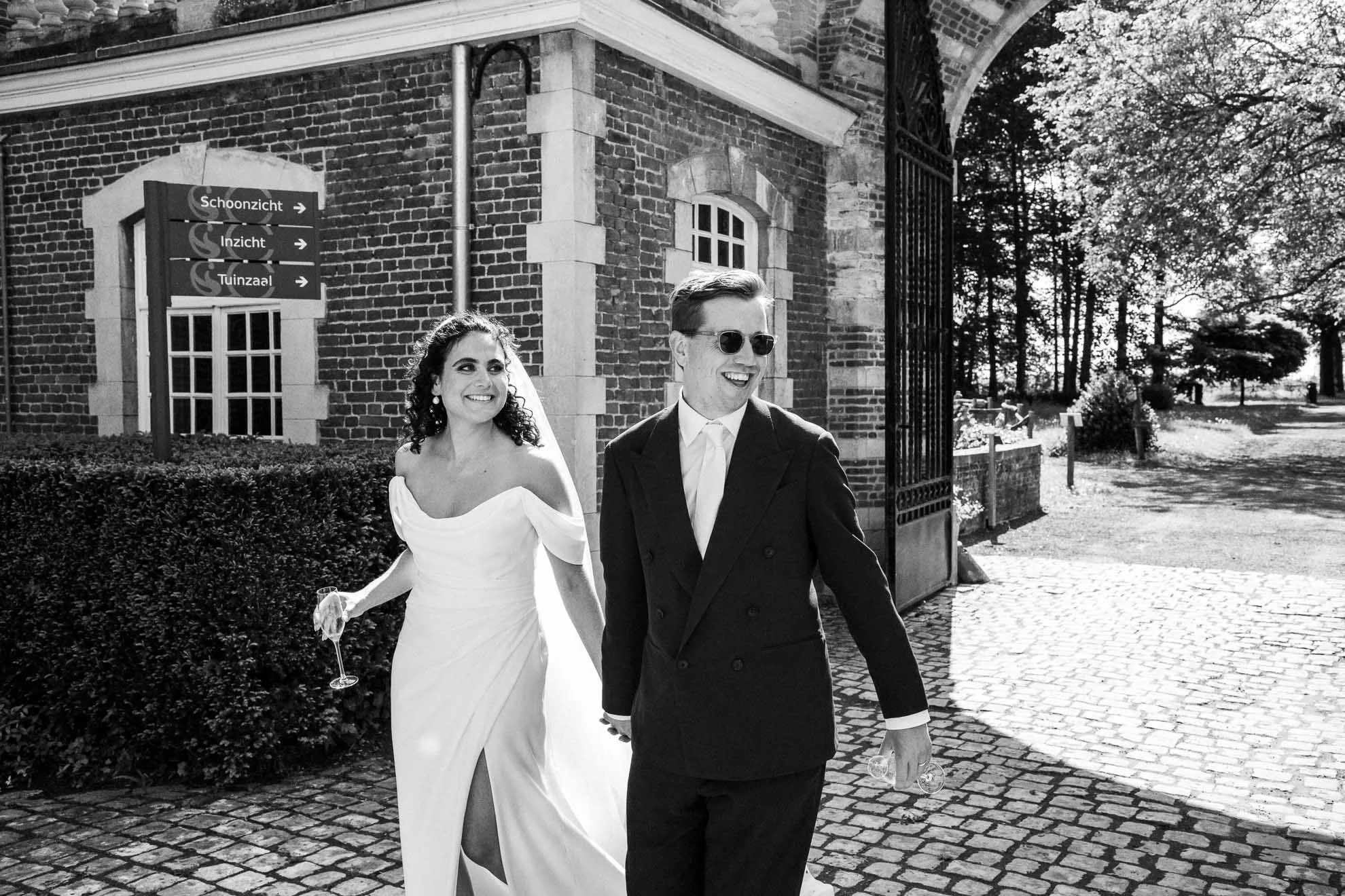 A smiling bride and groom walking outdoors near a brick building with a gate, trees, and path in the background. The bride is wearing a white wedding dress and holding a glass of champagne, while the groom is dressed in a dark suit and sunglasses.