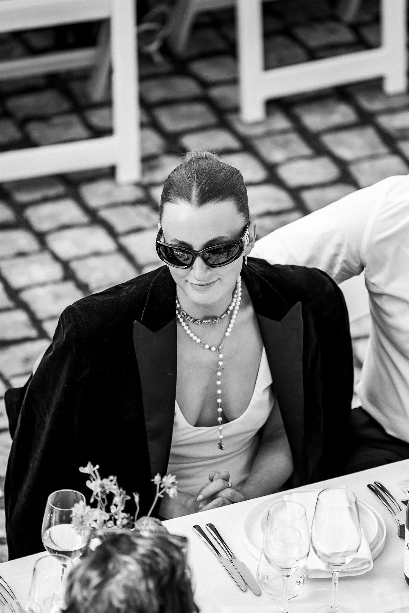 A woman in sunglasses, pearl jewelry, and a blazer, seated at a table with wine glasses and a floral centerpiece, smiling at a formal event.