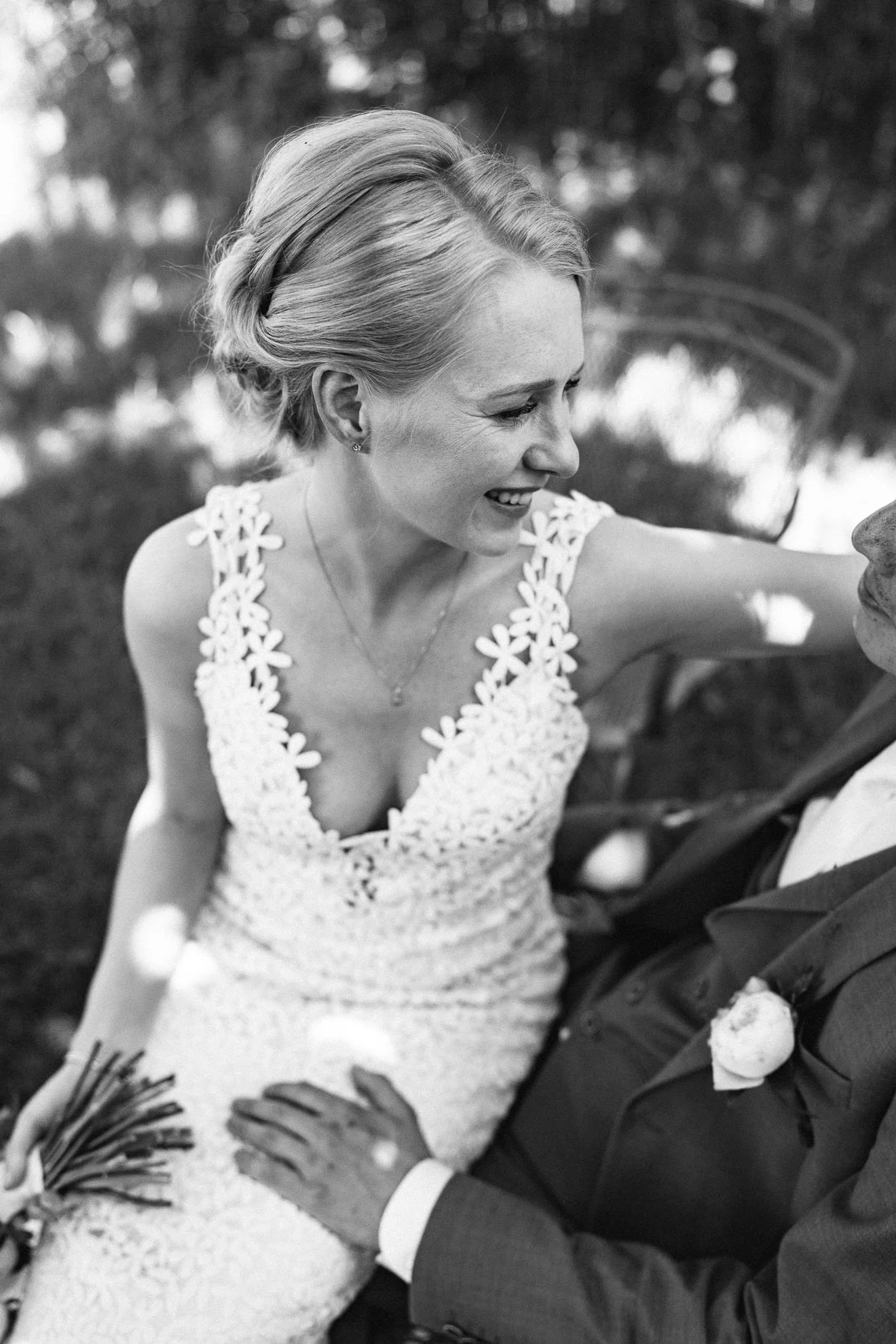 A black and white photo of a smiling woman in a wedding dress, looking at her groom, whose hands are resting on her waist. The woman has short, styled hair and is holding a bouquet. The background is blurred with nature scenery.
